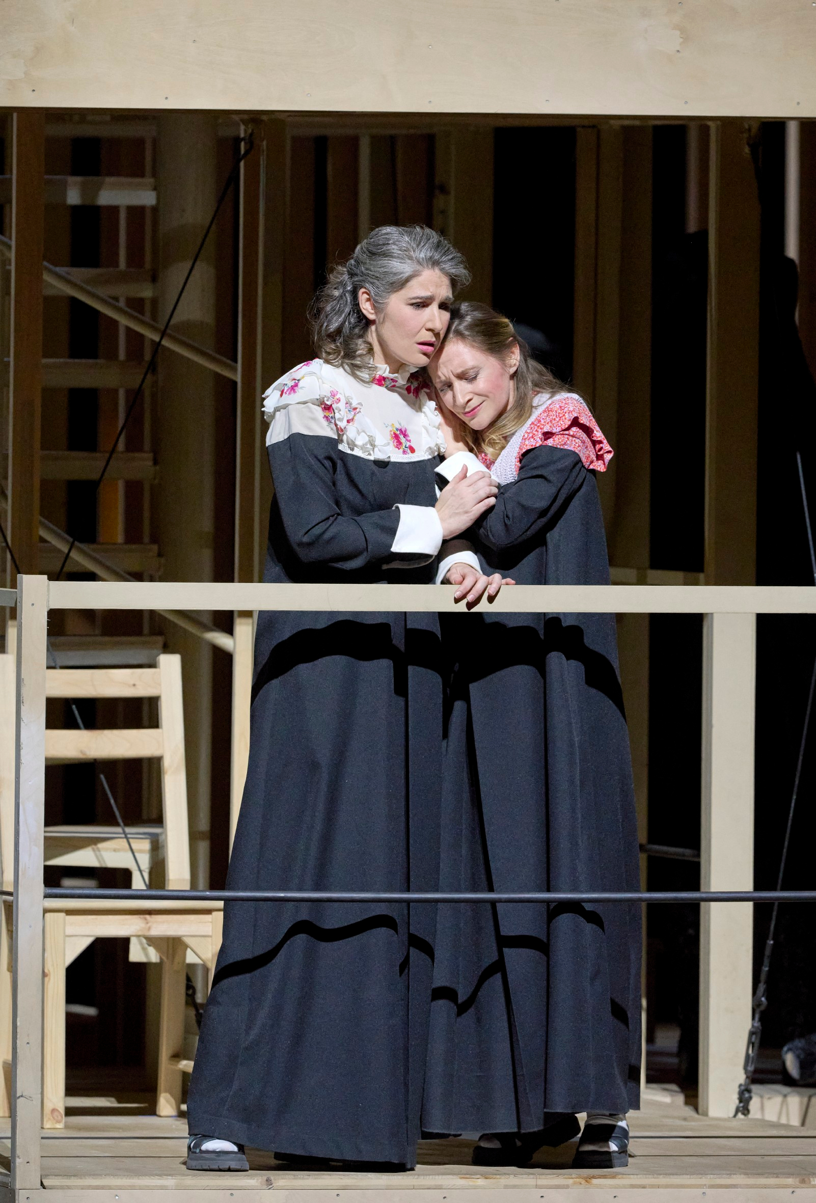 Zwei Frauen in dunkler Kleidung mit besticktem Kragen stehen eng umarmt an einem hölzernen Geländer und wirken emotional bewegt.