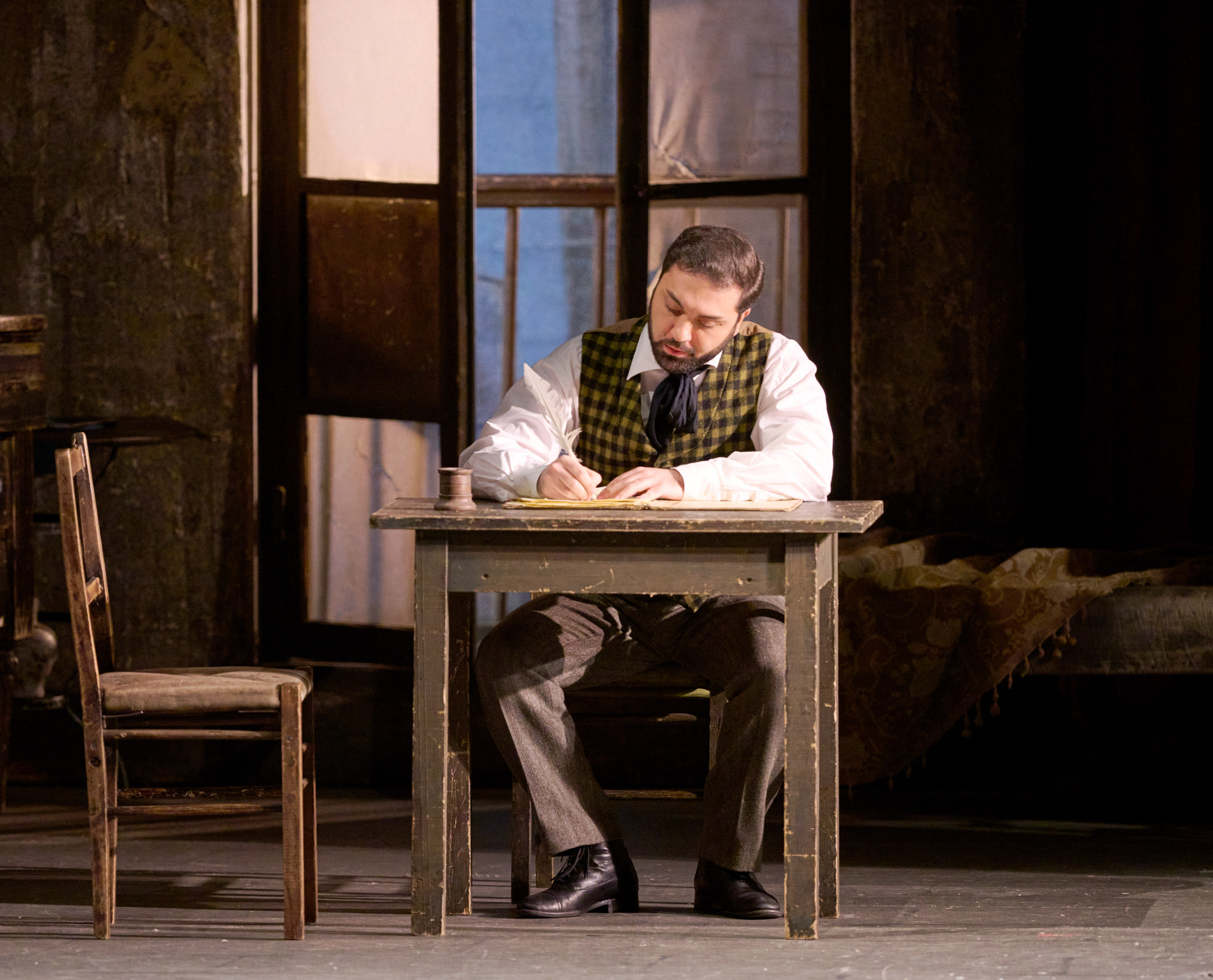 Ein Mann sitzt an einem schlichten Holztisch und schreibt konzentriert, im Hintergrund ein geöffnetes Fenster mit Vorhang.