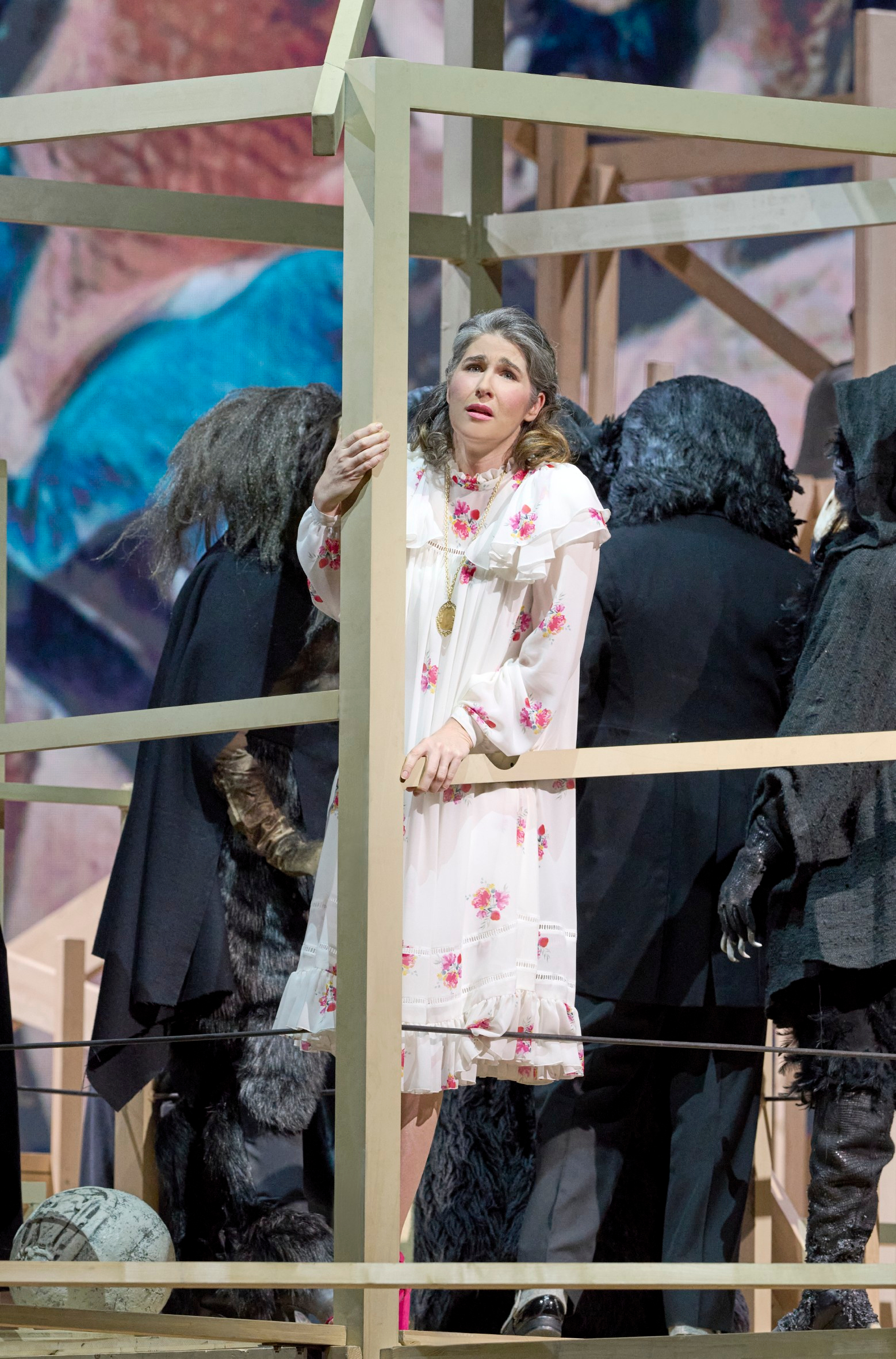 Eine Frau im weißen Nachthemd mit Blumenmuster steht ängstlich in einem Holzrahmen, umgeben von dunkel gekleideten Gestalten mit schwarzen Perücken.