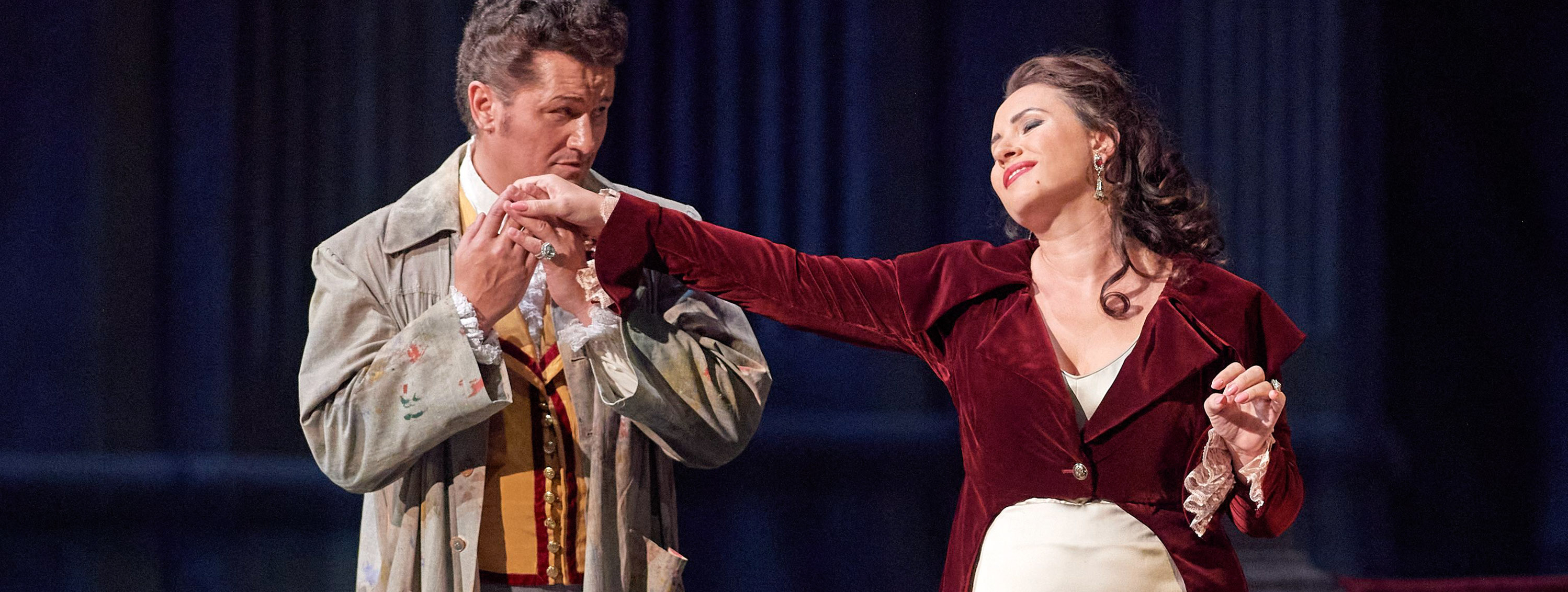 Ein Mann küsst die Hand einer Frau, die in einem roten Samtmantel und einem eleganten weißen Kleid vor ihm steht, während sie ihm liebevoll in die Augen blickt.