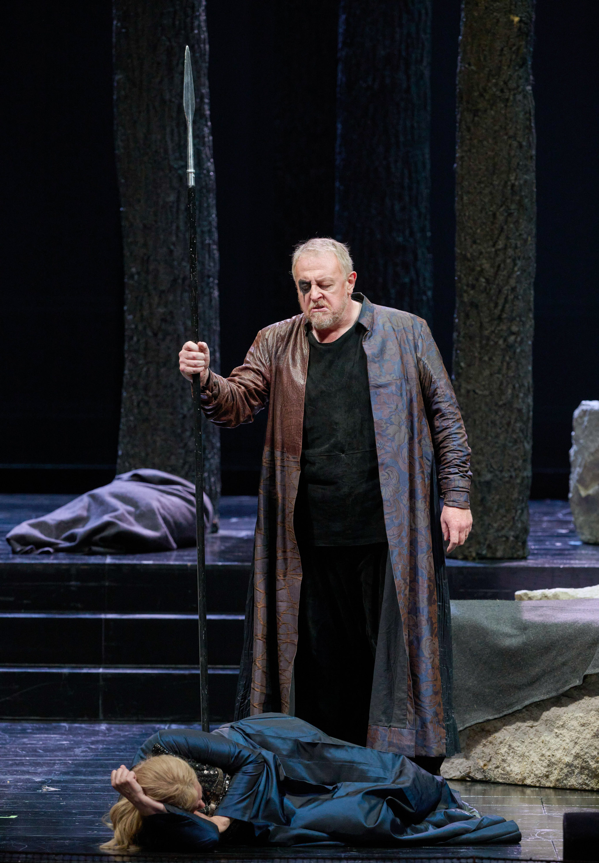 Ein Mann in langem Mantel hält einen Stab auf einer dunklen Bühne mit steinernen Elementen.