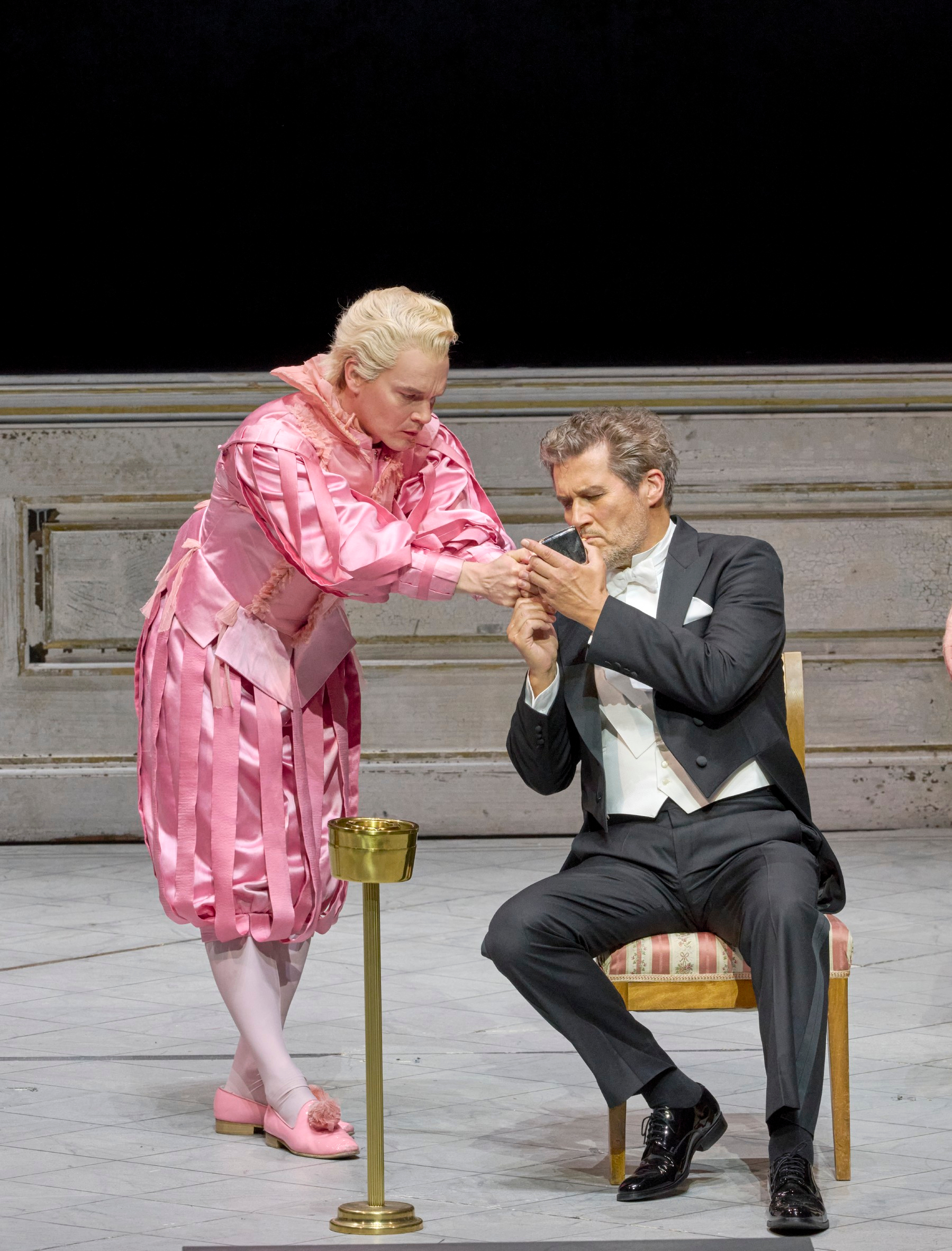 Eine Person in auffälligem rosa Kostüm reicht einem Mann im Smoking etwas, während dieser aufmerksam hinsieht.