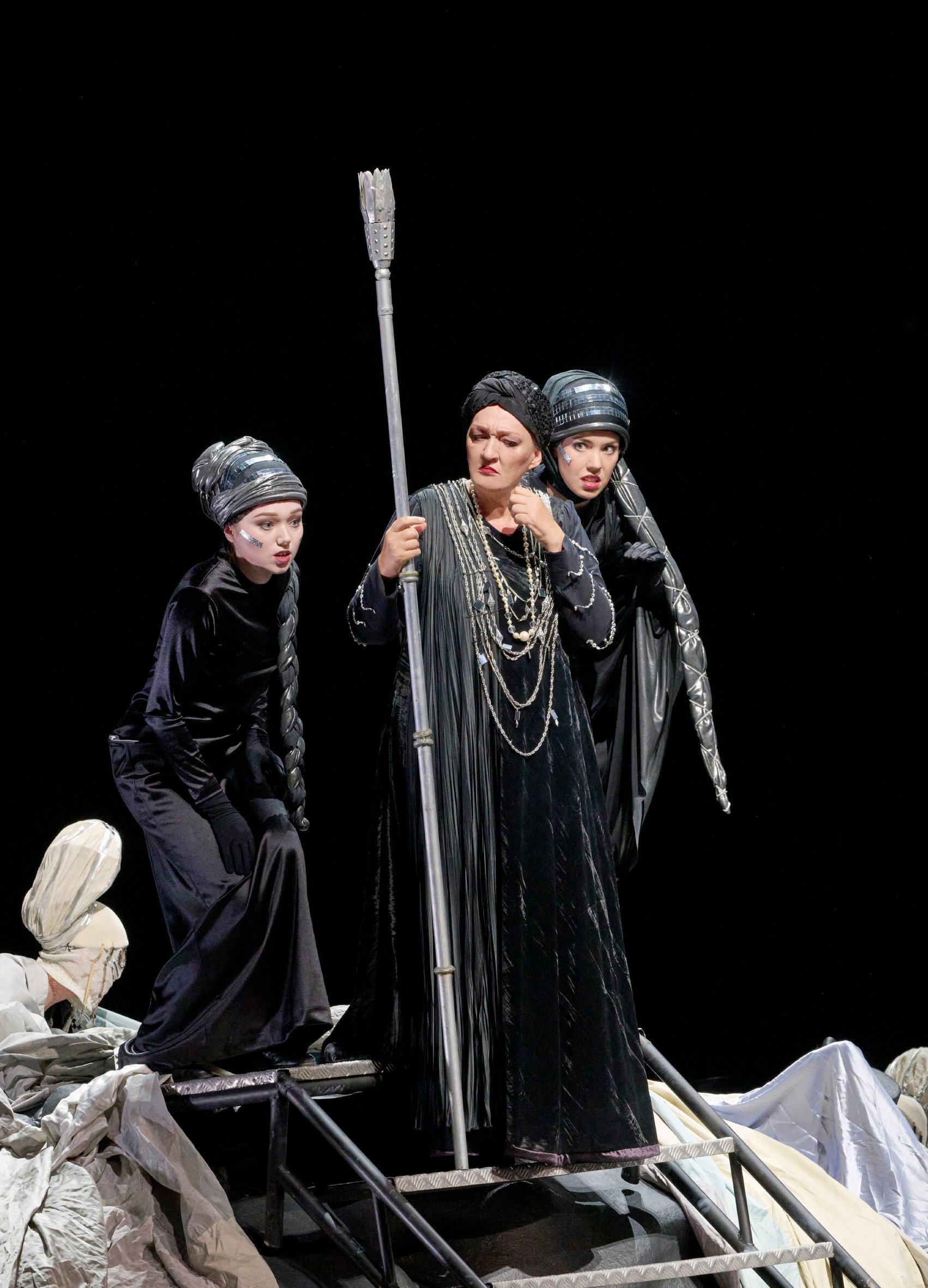 Drei Darstellerinnen in dunklen Kostümen stehen auf der Bühne, eine Person hält einen langen Stab und trägt auffälligen Kopfschmuck.