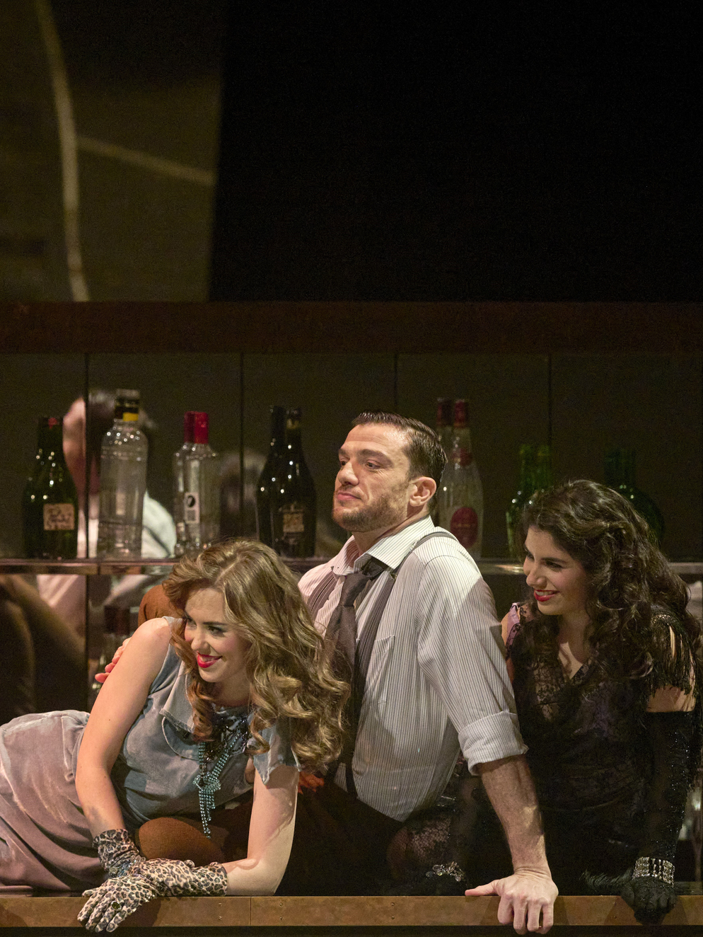 Ein Mann steht lässig an einer Bar, flankiert von zwei Frauen in aufreizender Kleidung, mit Flaschen im Hintergrund.