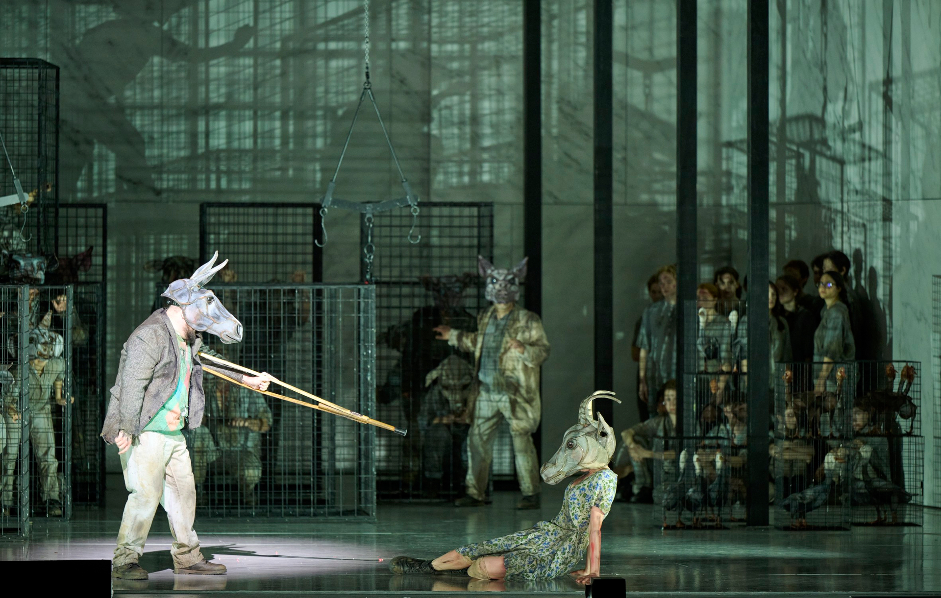 Eine Figur mit Tiermaske zielt mit einer langen Waffe auf eine am Boden liegende Person mit Ziegenmaske, während andere in Käfigen im Hintergrund zusehen.