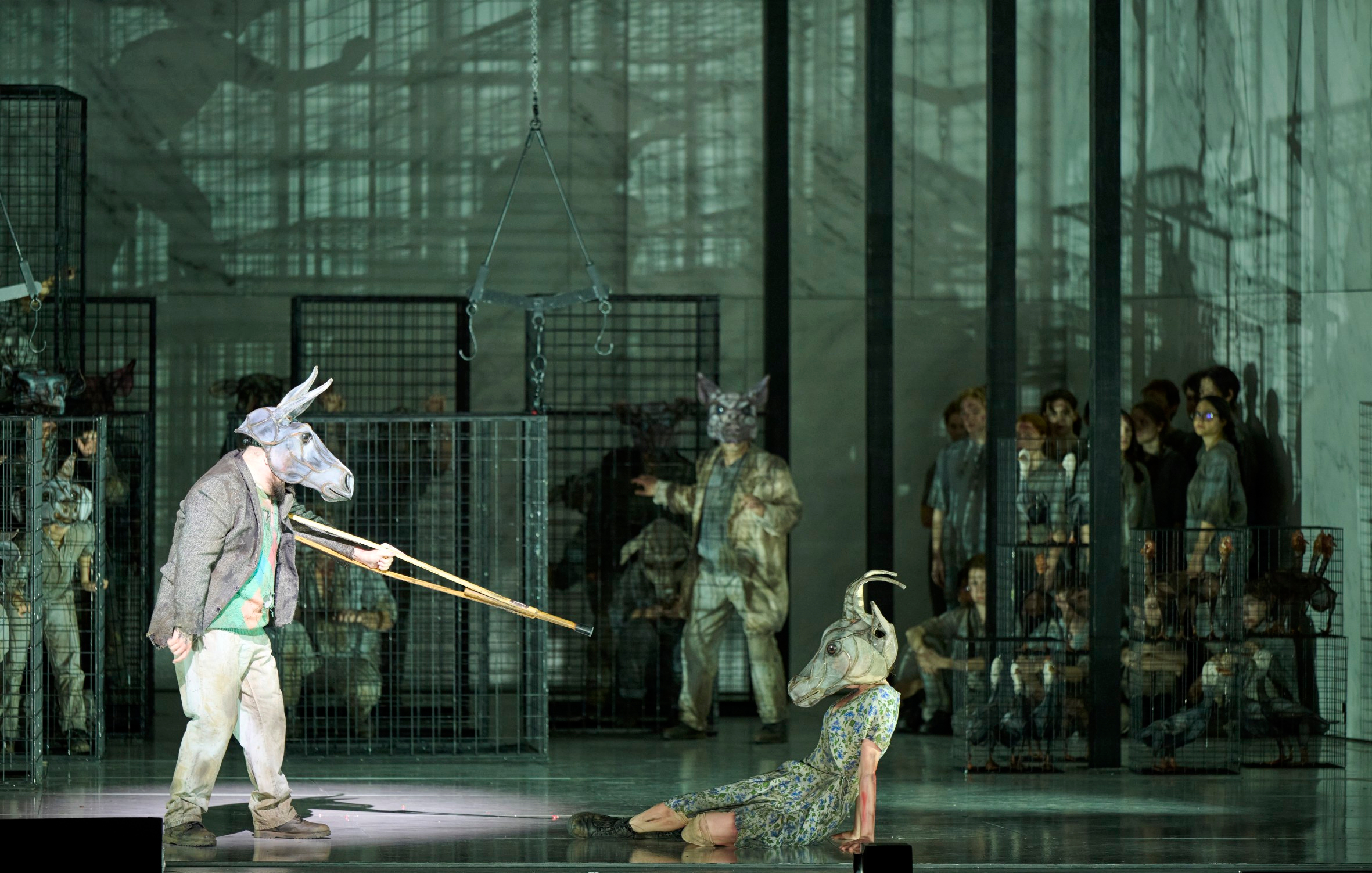 Eine Figur mit Tiermaske zielt mit einer langen Waffe auf eine am Boden liegende Person mit Ziegenmaske, während andere in Käfigen im Hintergrund zusehen.