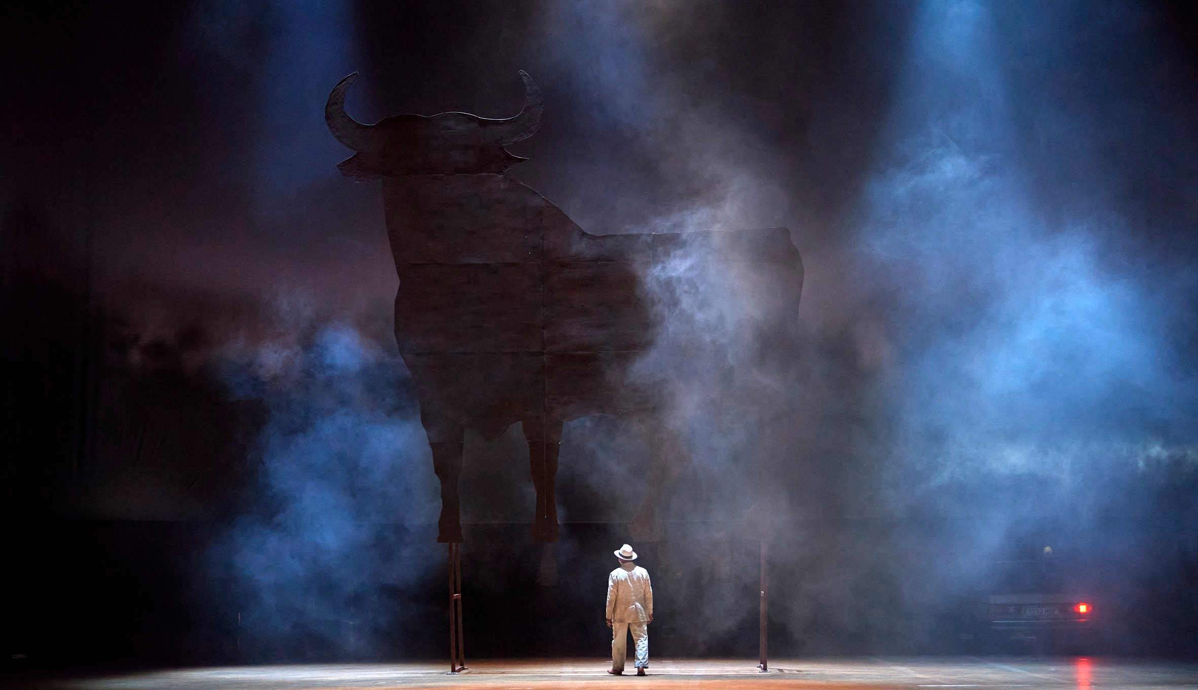 Eine einzelne Figur in heller Kleidung steht vor einer riesigen schwarzen Silhouette eines Stiers, umgeben von dichtem Nebel auf der Bühne.