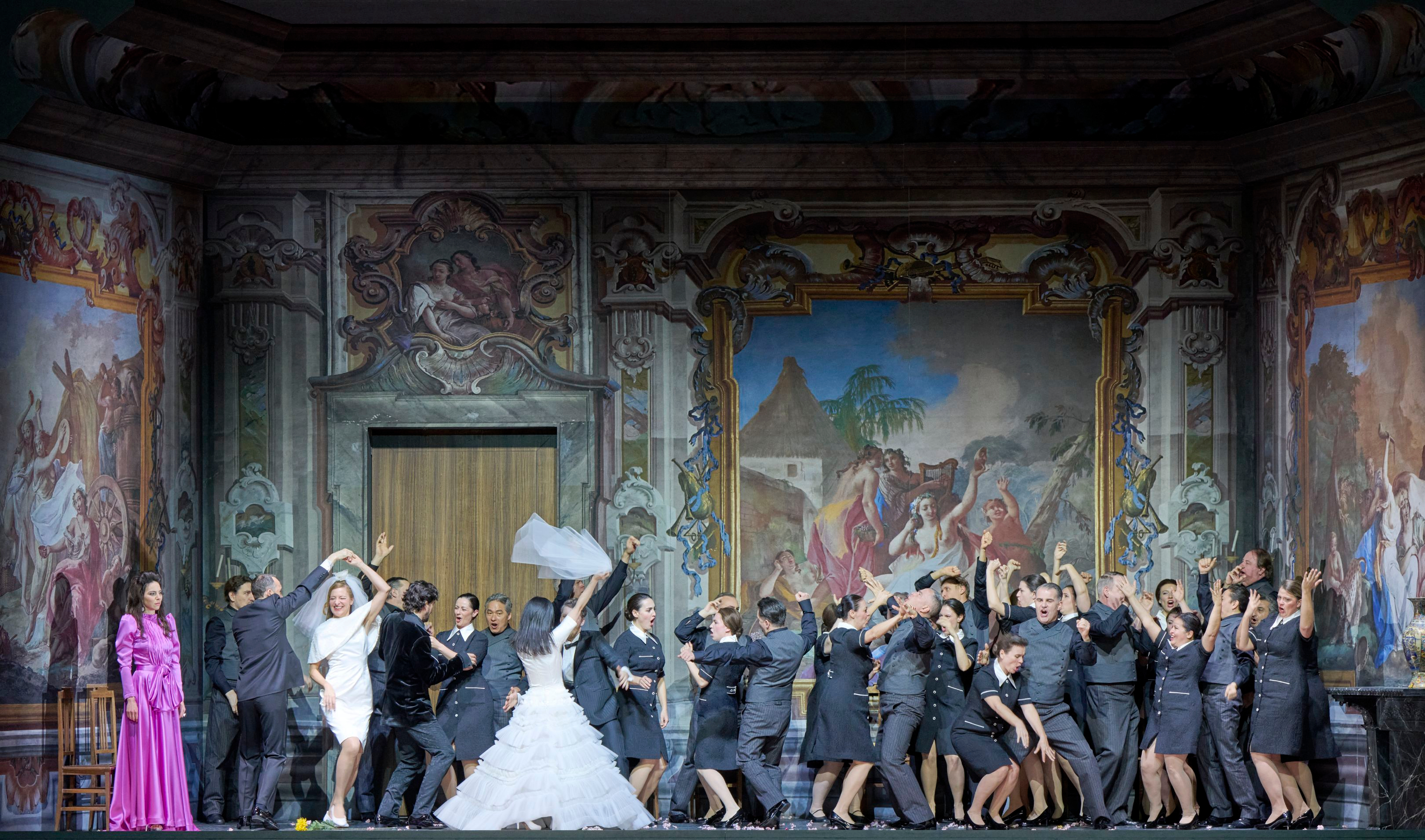 Vor einer prunkvoll bemalten Wand tanzt eine große Gruppe in grauen Anzügen, während eine Frau im weißen Brautkleid jubelnd den Schleier schwingt.