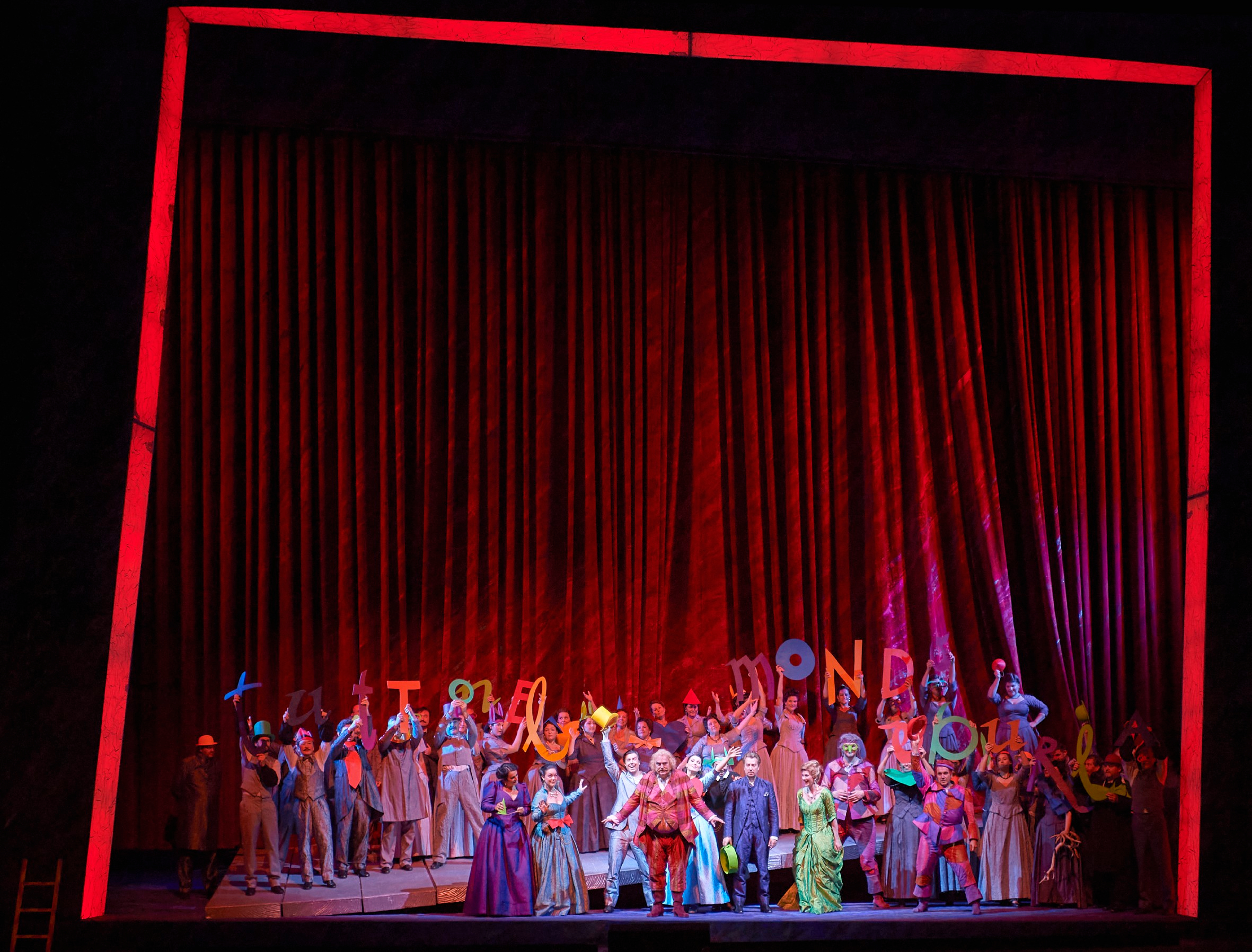 Eine große Gruppe bunt gekleideter Menschen steht jubelnd auf der Bühne vor einem geschlossenen roten Vorhang und hält Buchstaben hoch.