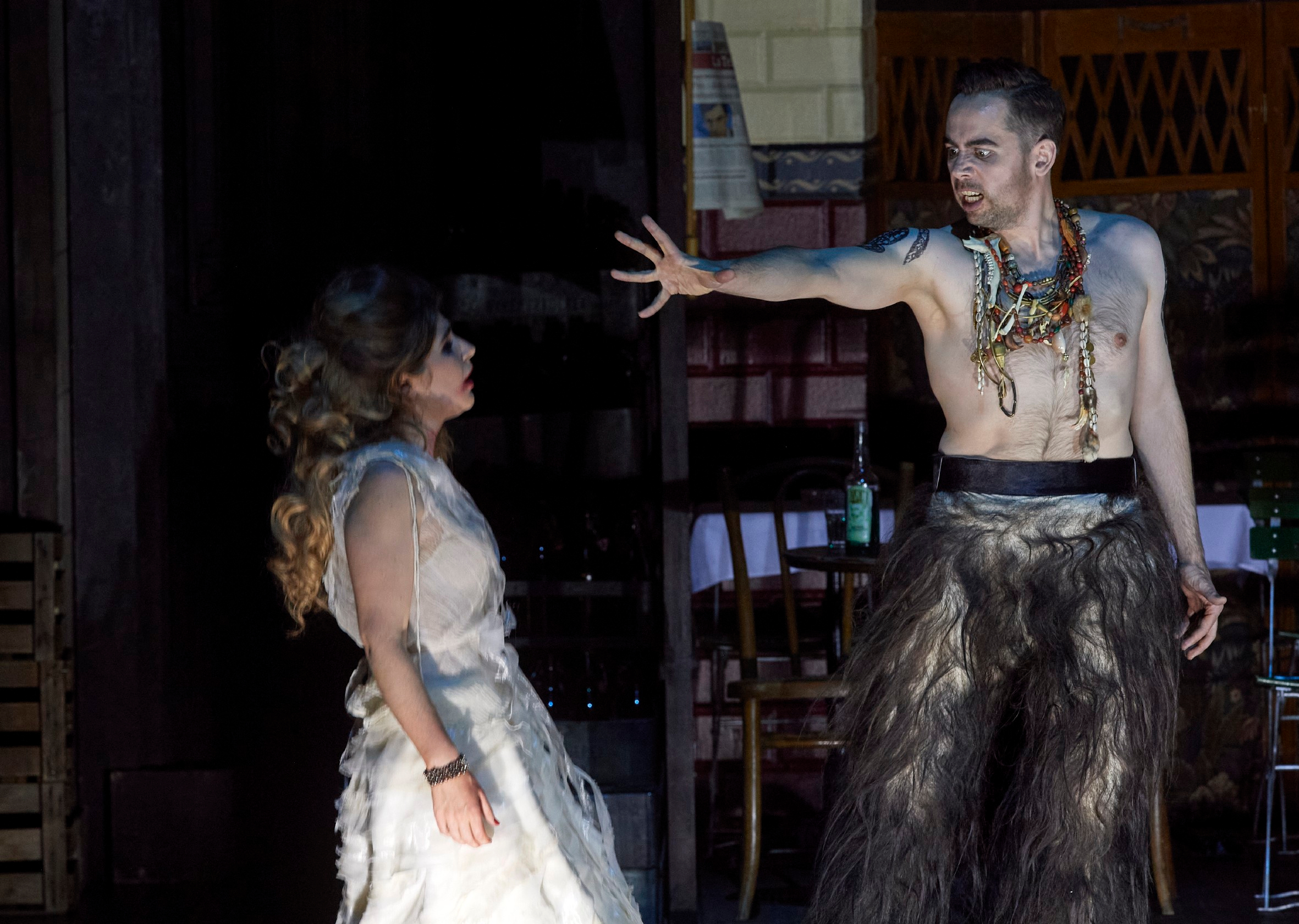 Ein Mann in wildem, tänzerischem Kostüm streckt dramatisch den Arm aus, während eine Frau in weißem Kleid ihn anschaut.