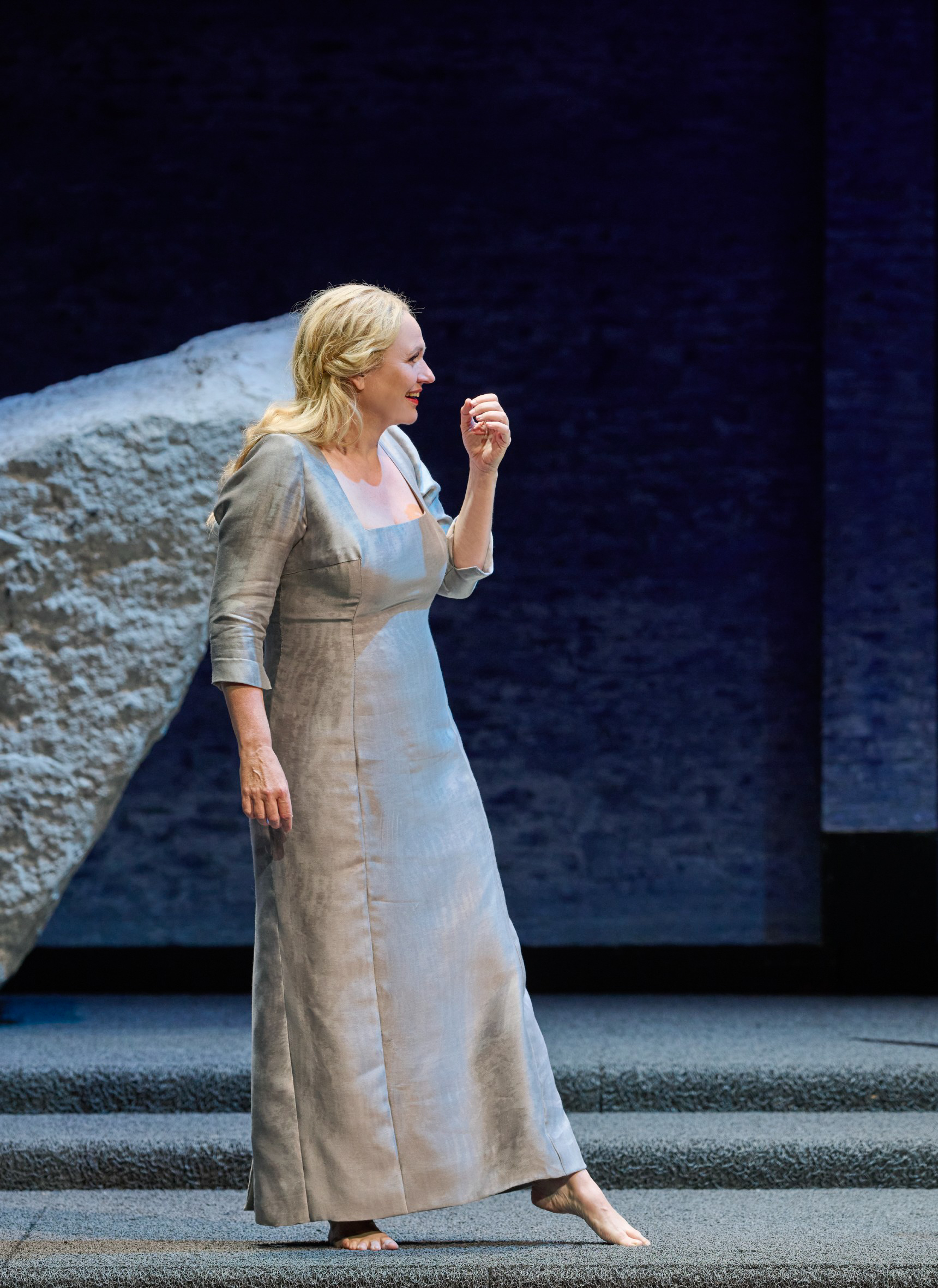 Eine Frau in hellem Kleid steht vor einem Felsen und blickt zur Seite.