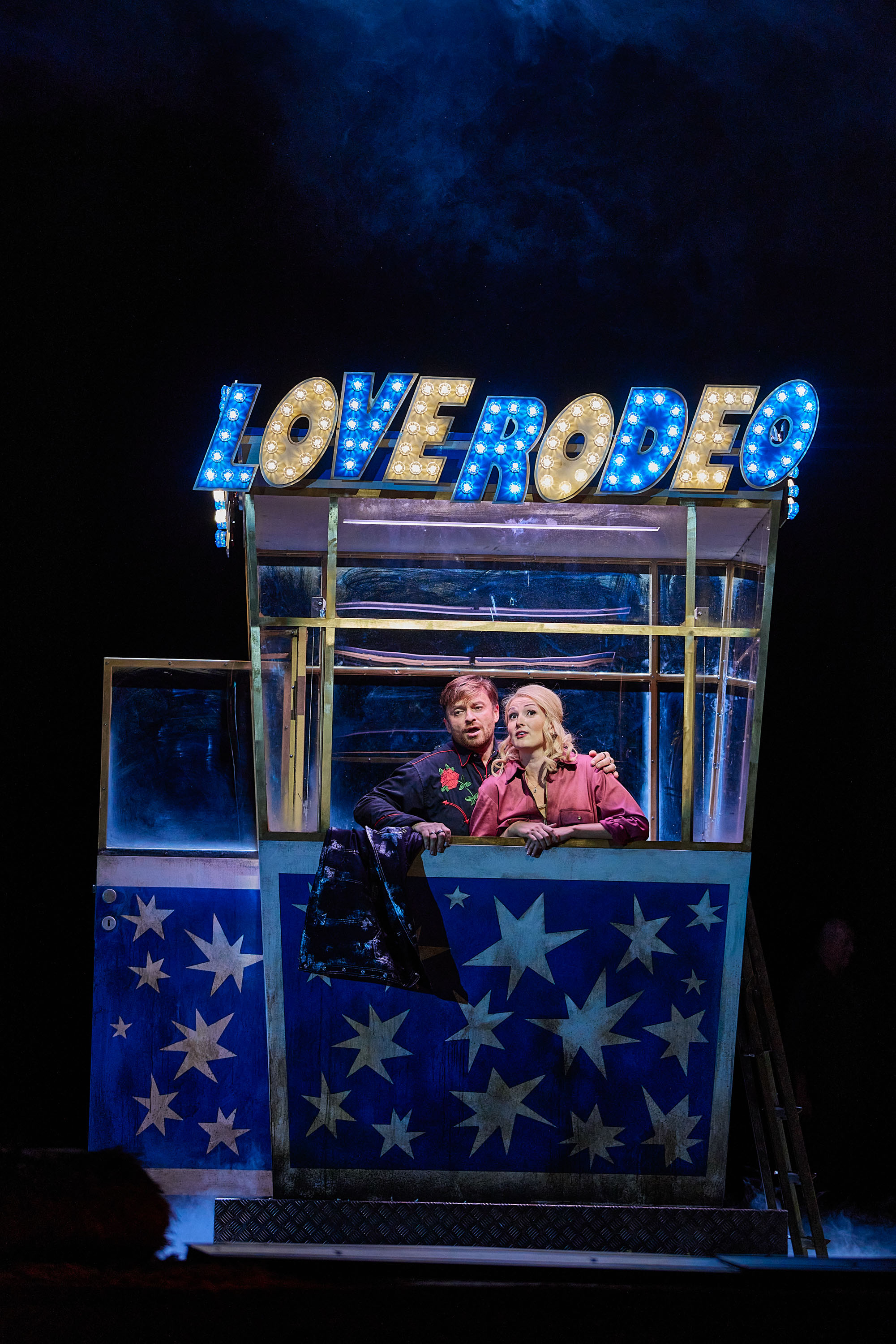 Ein Mann und eine Frau stehen gemeinsam in einer kleinen blauen Kabine mit weißen Sternen darauf. Am Dach ist der Schriftzug "Love Rodeo" angebracht.