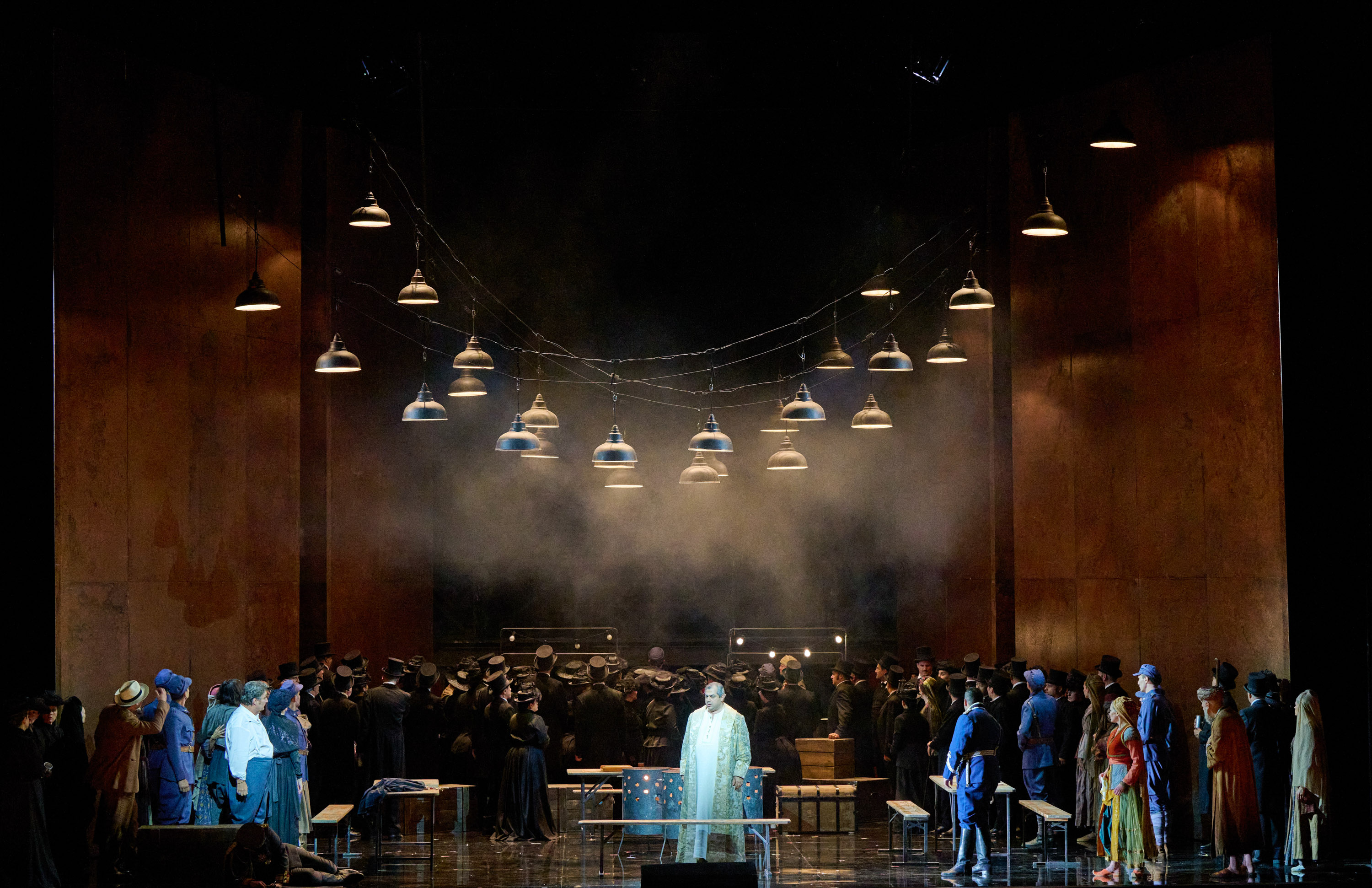 Eine Gruppe Menschen steht auf der Bühne. Über ihnen hängen viele Lampen. Im Zentrum steht ein Mann in einem langen weißen Gewand.