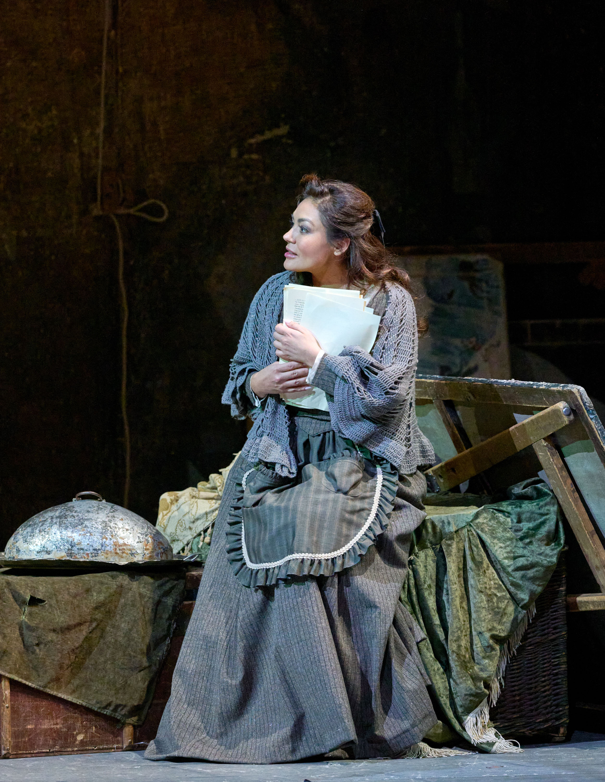 Eine Frau in schlichter Kleidung sitzt auf einer Kiste und drückt einen Brief fest an sich, während sie versonnen zur Seite blickt.