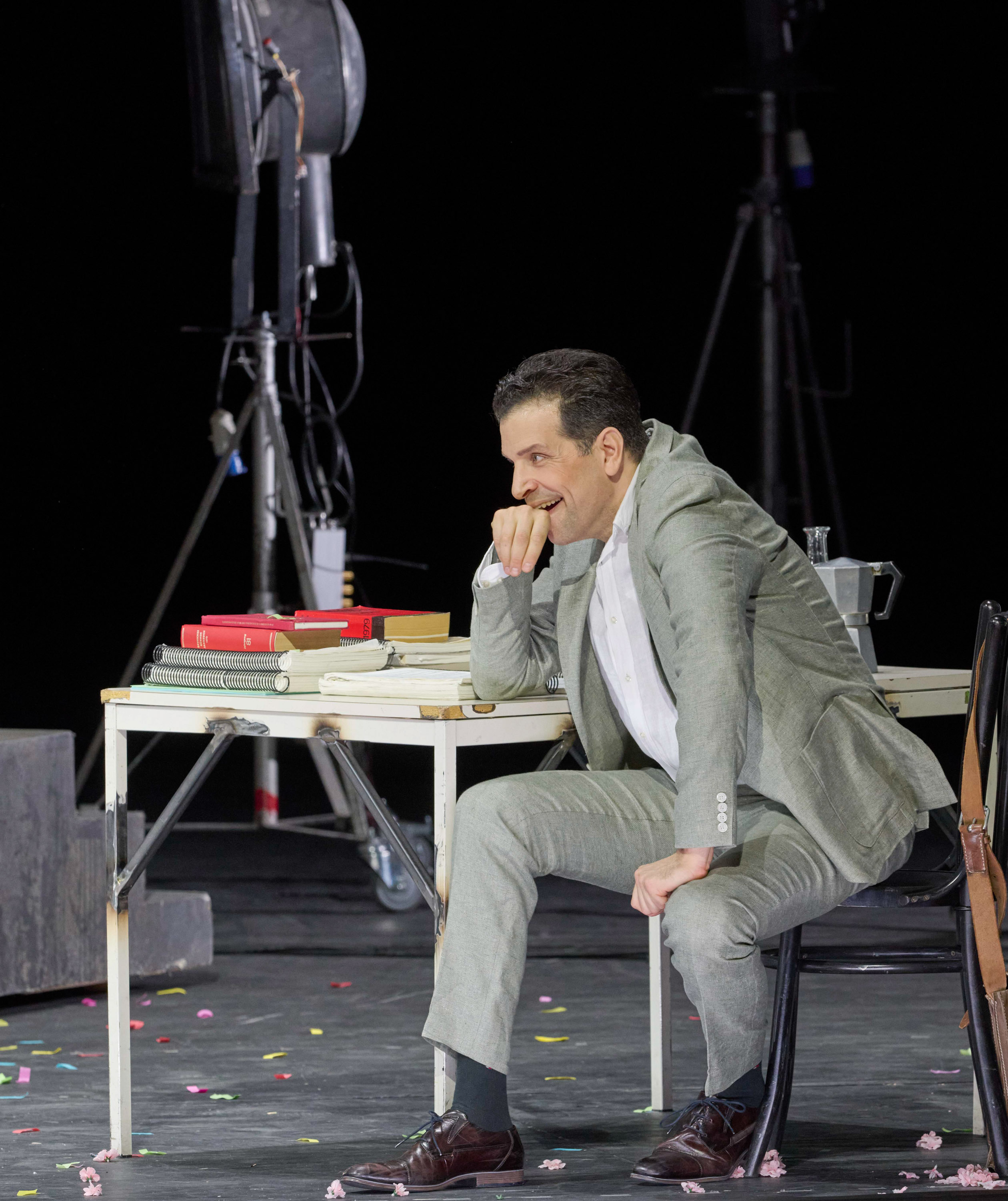 Ein Mann in einem hellgrauen Anzug sitzt nachdenklich an einem Tisch mit Büchern und einem Wasserkrug, umgeben von Bühnenbeleuchtung und Kameraequipment.