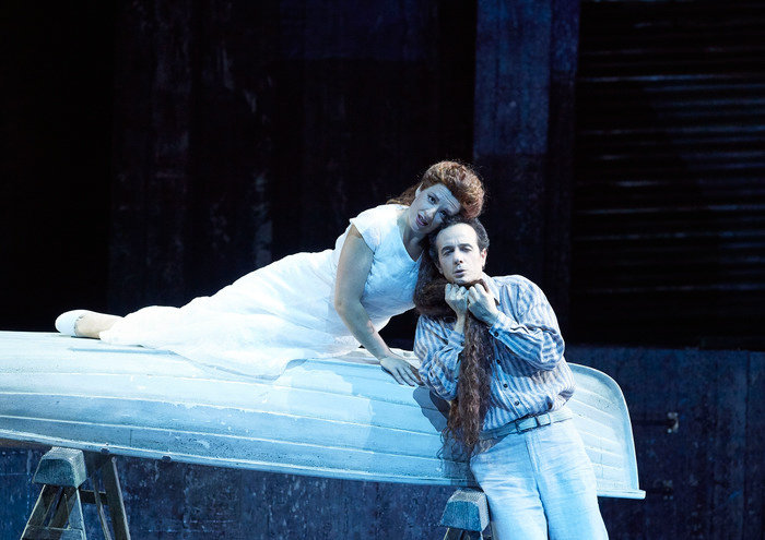 Eine Frau im weißen Kleid liegt auf einem Bett und beugt sich zu einem Mann, der zärtlich ihr langes Haar hält.