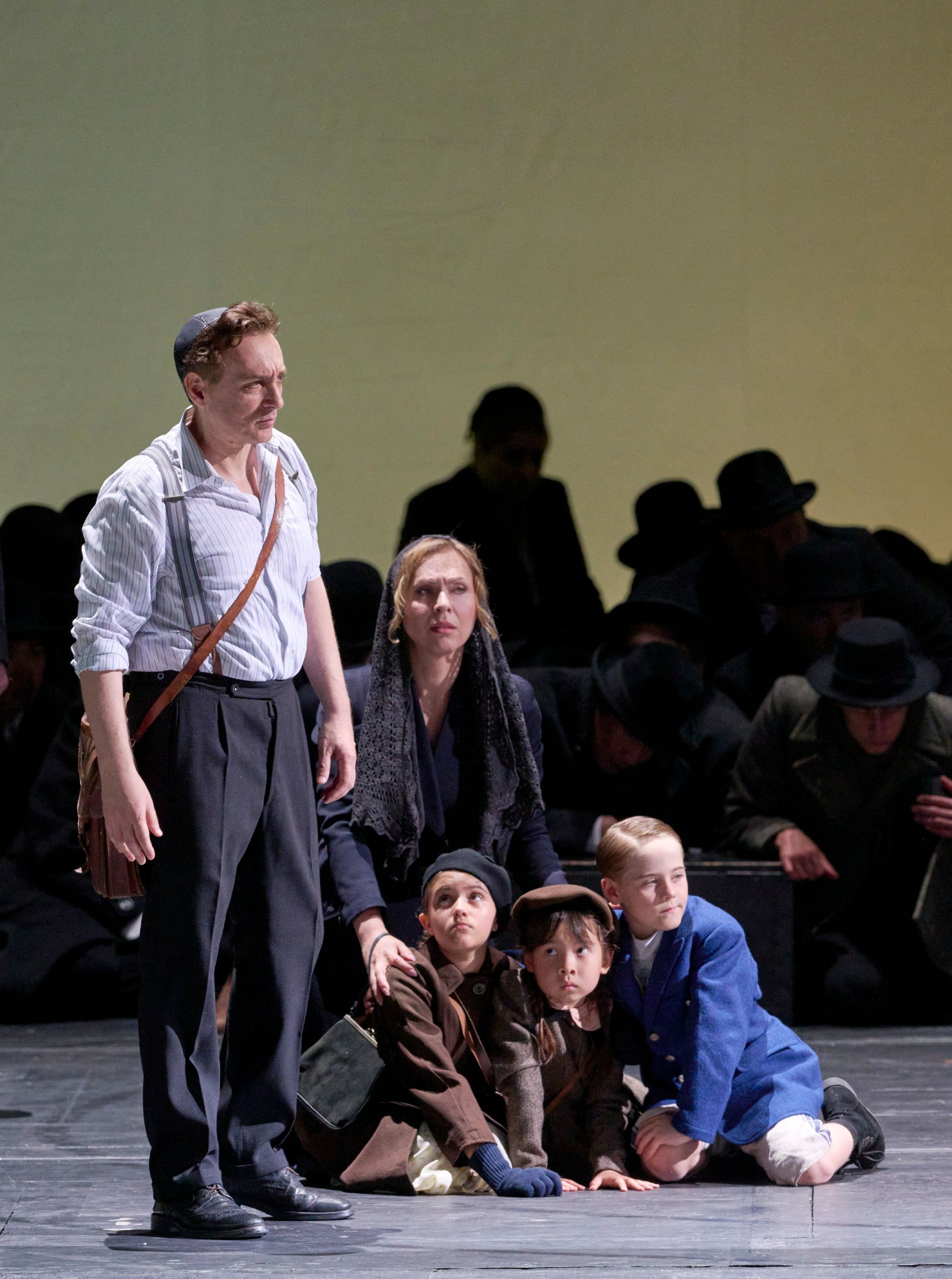 Ein Mann steht angespannt neben einer Gruppe von Frauen und Kindern am Boden, umgeben von dunkel gekleideten Gestalten im Hintergrund.