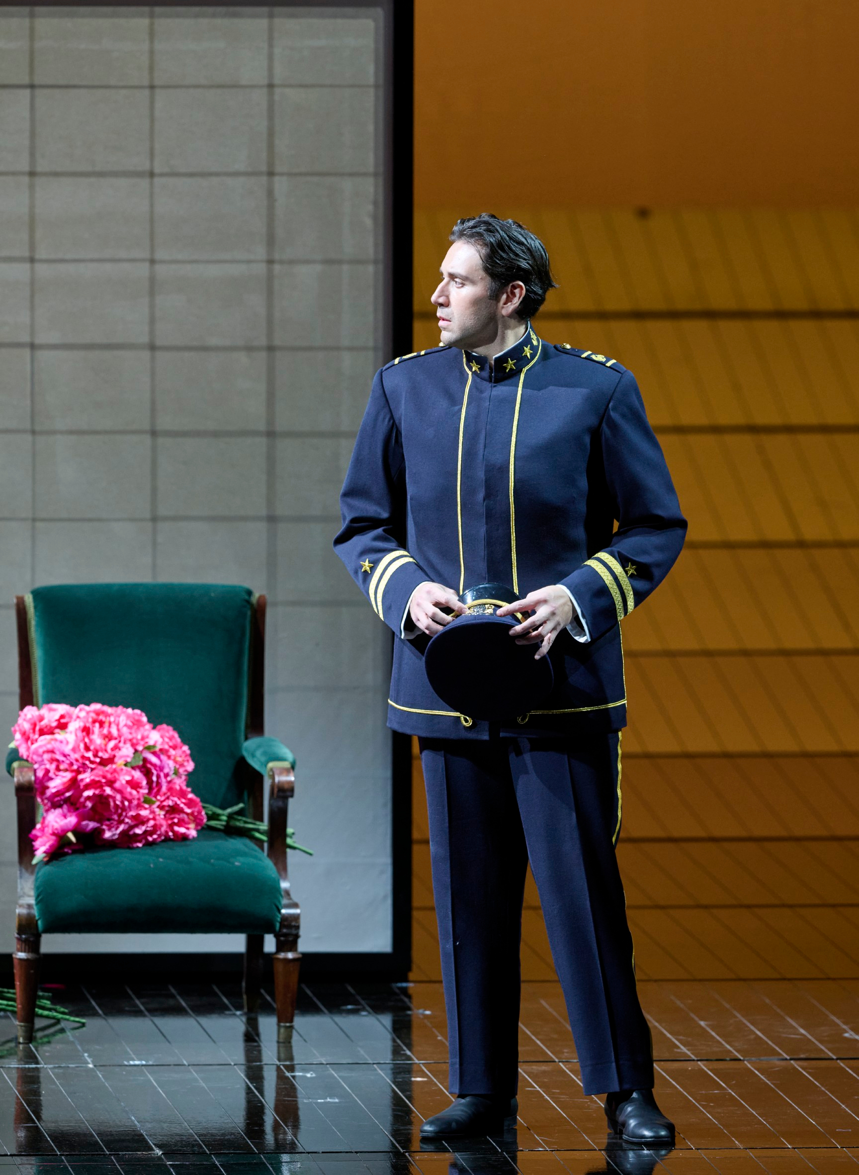 Ein Mann in dunkelblauer Uniform steht mit Mütze in der Hand neben einem grünen Sessel mit einem Strauß pinker Rosen.