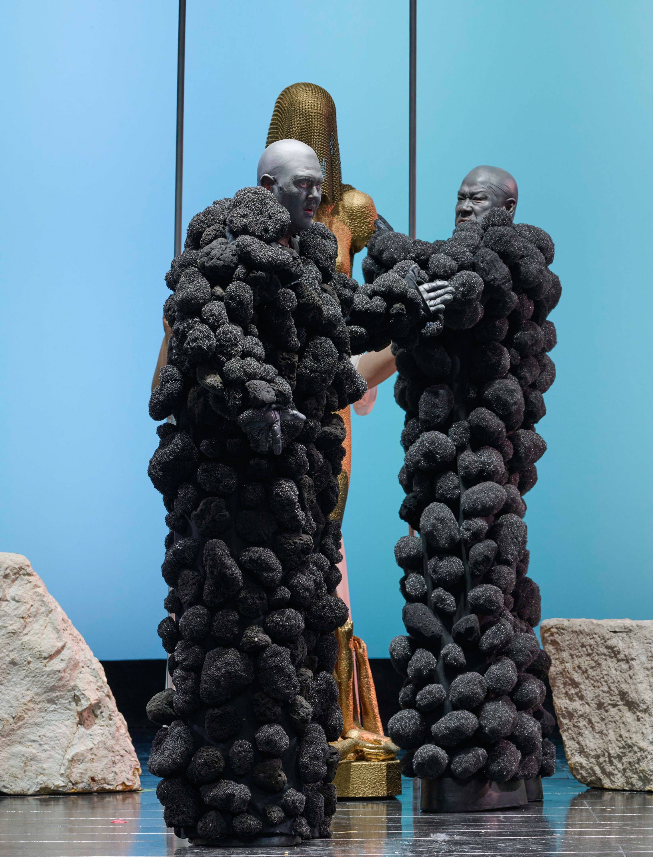 Zwei dunkel-felsenartig kostümierte Männer stehen vor einer goldenen Figur.