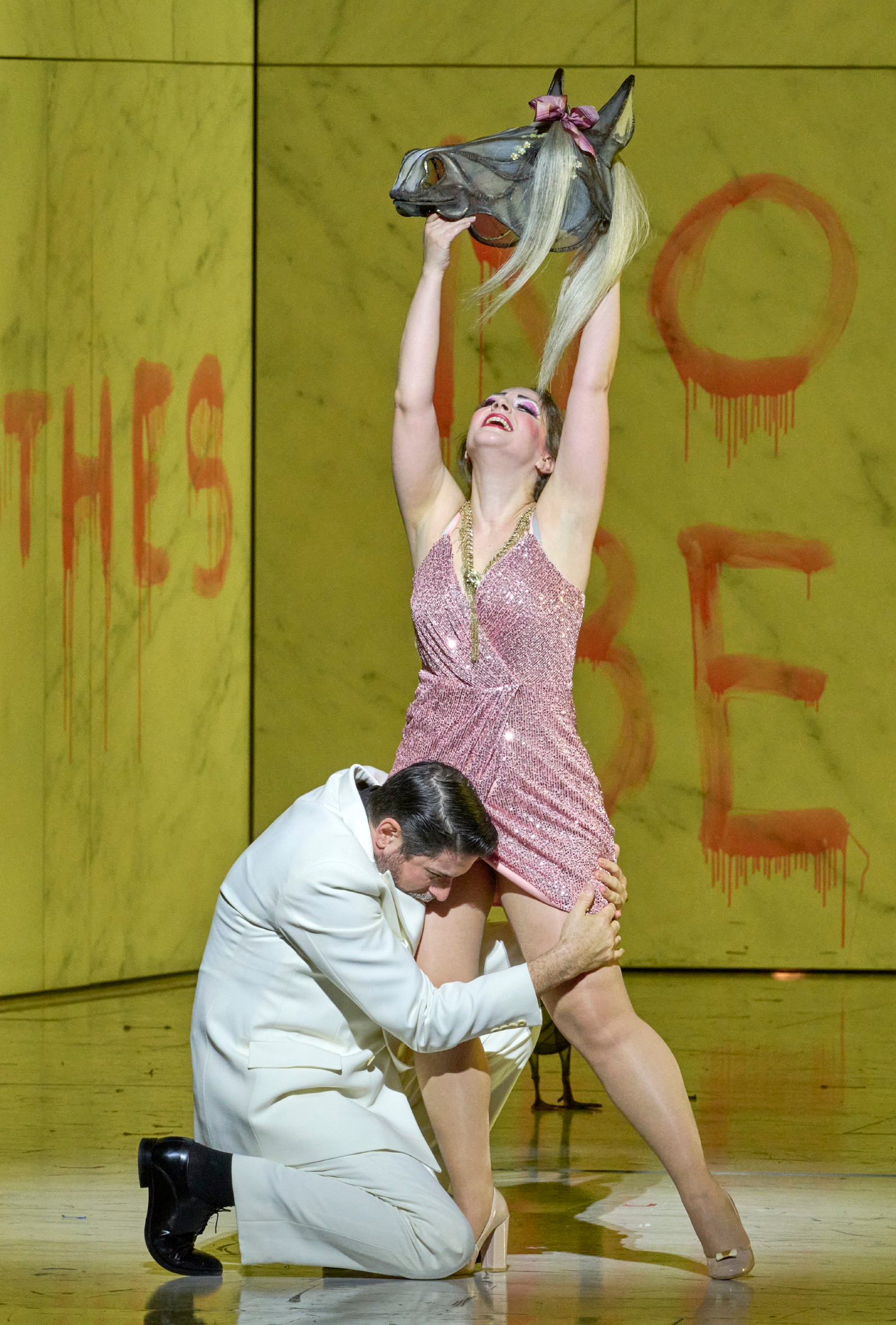 Eine Frau in glitzerndem Kleid hebt triumphierend eine Pferdemaske über den Kopf, während ein Mann sich vor ihr niederbeugt; im Hintergrund rote Schrift an gelber Wand.