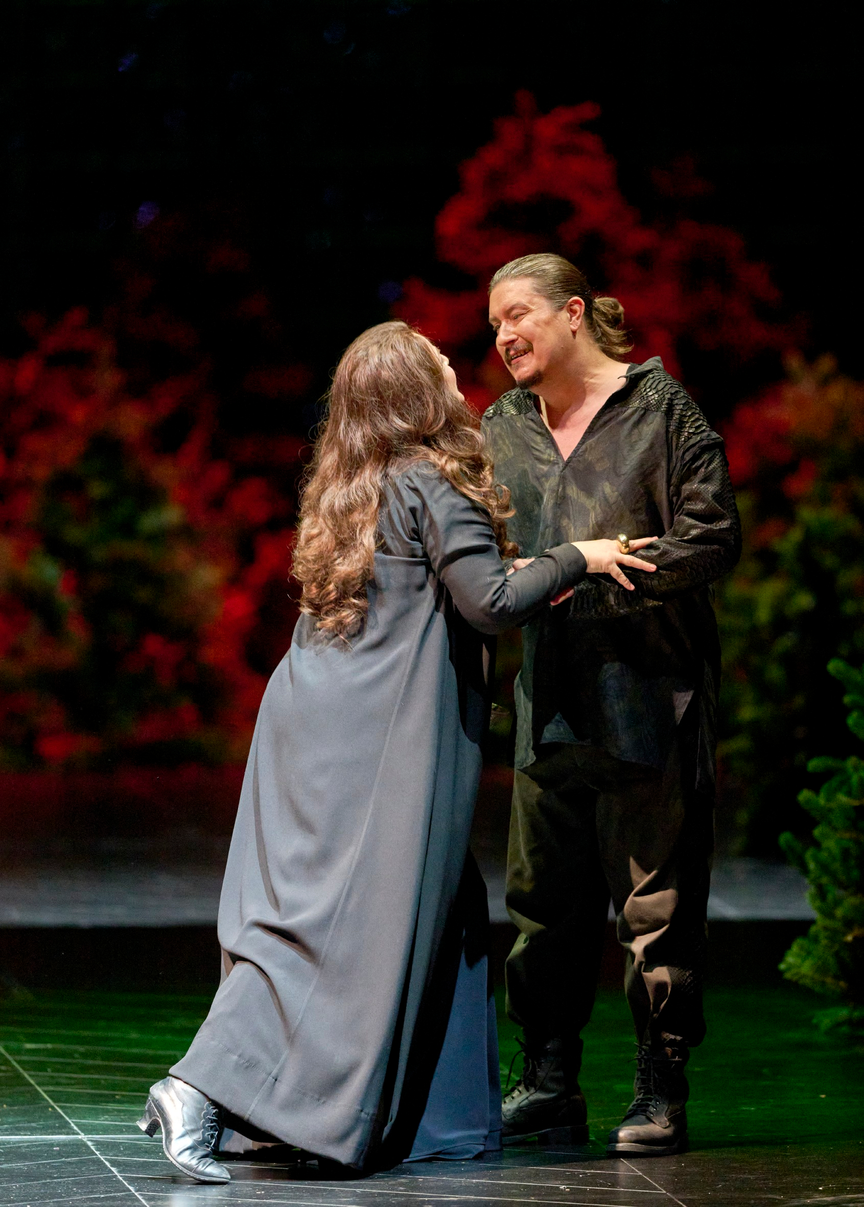 Zwei Personen stehen sich auf der Bühne vor einem roten Blätterhintergrund gegenüber, halten sich an den Händen und lächeln einander an.