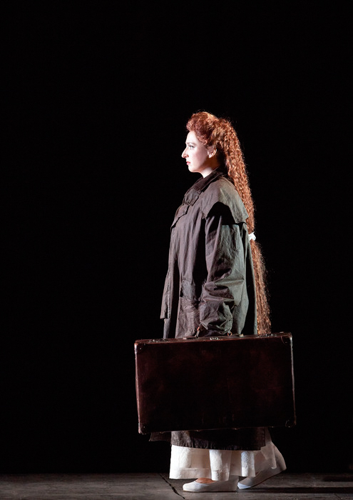 Eine Frau mit sehr langem, rotem Haar steht im Profil vor schwarzem Hintergrund und trägt eine dunkle Jacke.