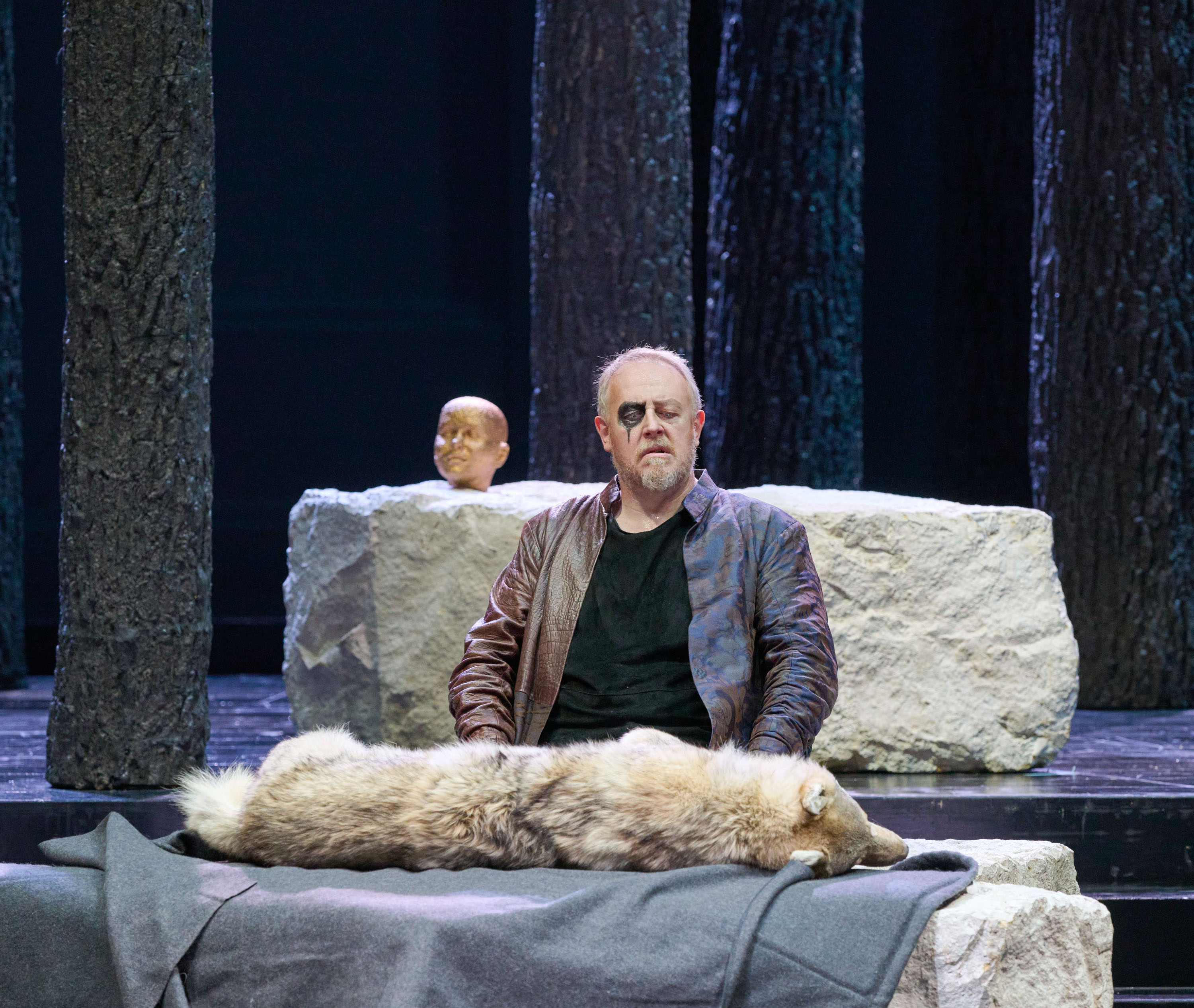 Ein Mann in Lederjacke sitzt auf einer Steinplatte mit einem Fell darauf, während im Hintergrund ein Kopfmodell auf einem Stein steht.