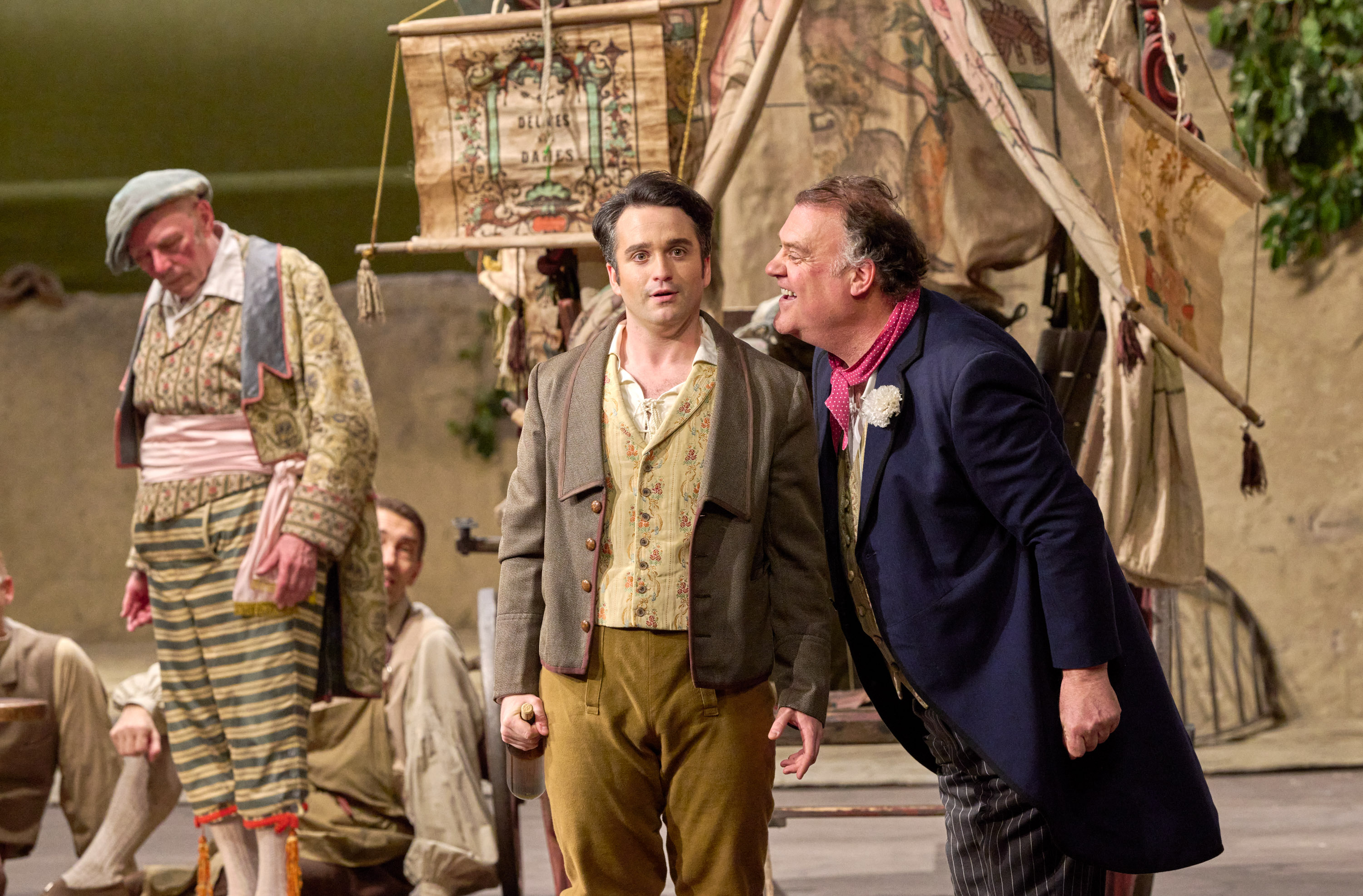 Ein älterer Mann beugt sich vor und spricht energisch mit einem jüngeren Mann, beide in historischer Kleidung vor einem dekorierten Zelt.