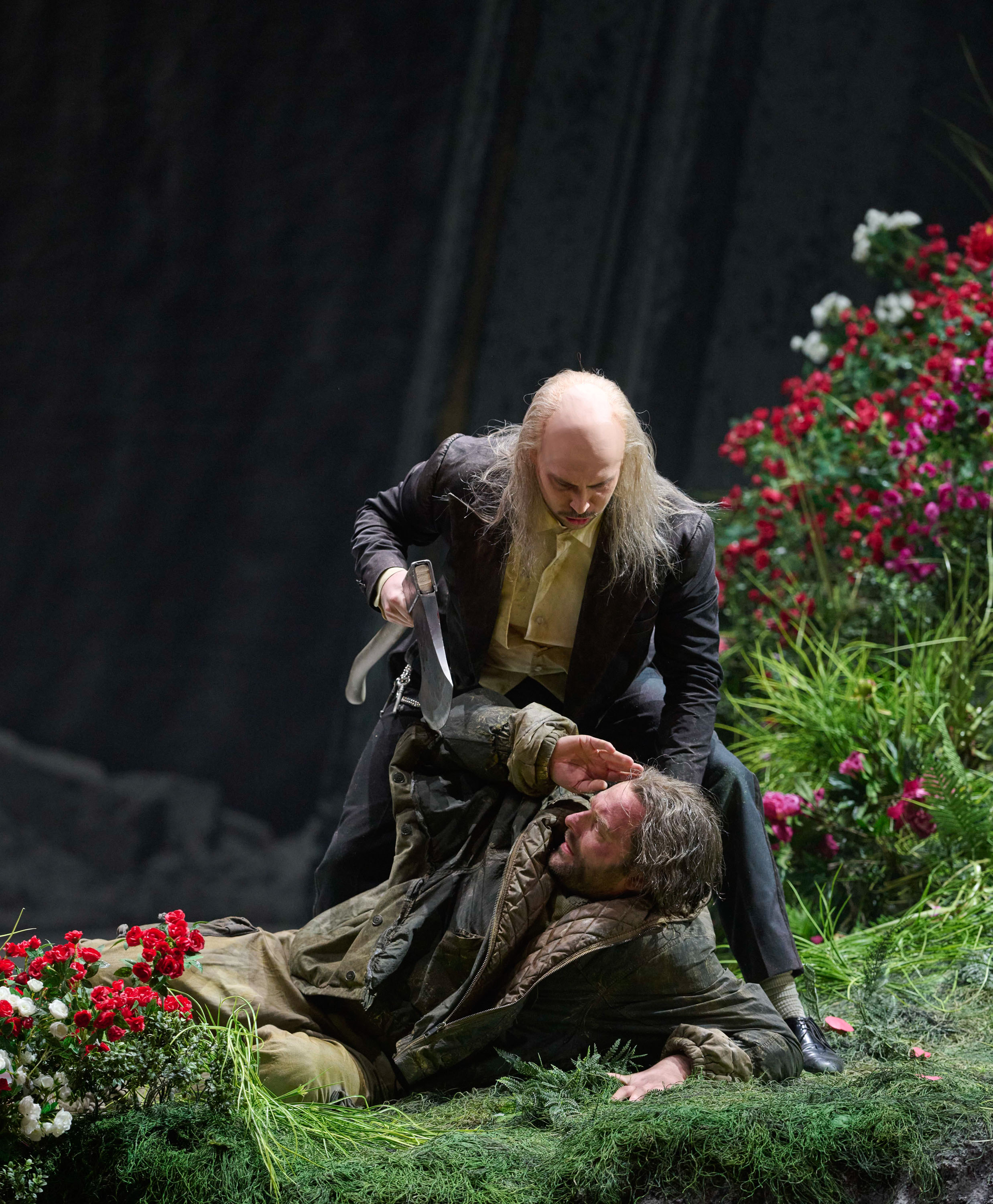 Ein älterer Mann mit Dolch bedroht einen am Boden liegenden Jüngeren auf einer blumenbewachsenen Bühne.