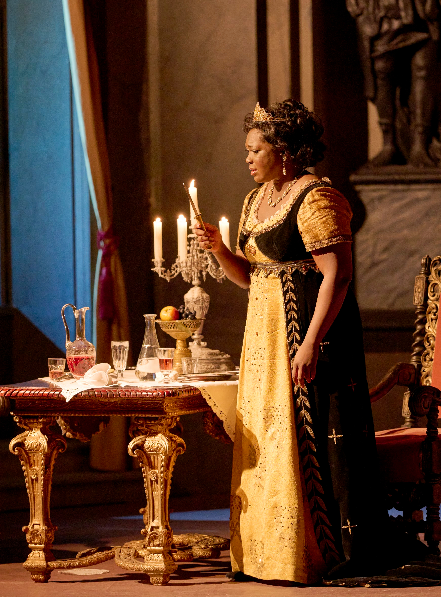 Eine Frau in einem eleganten gelben Kleid hält eine Kerzenleuchter vor einem Tisch, auf dem Getränke und Früchte stehen, in einem opulent eingerichteten Raum.