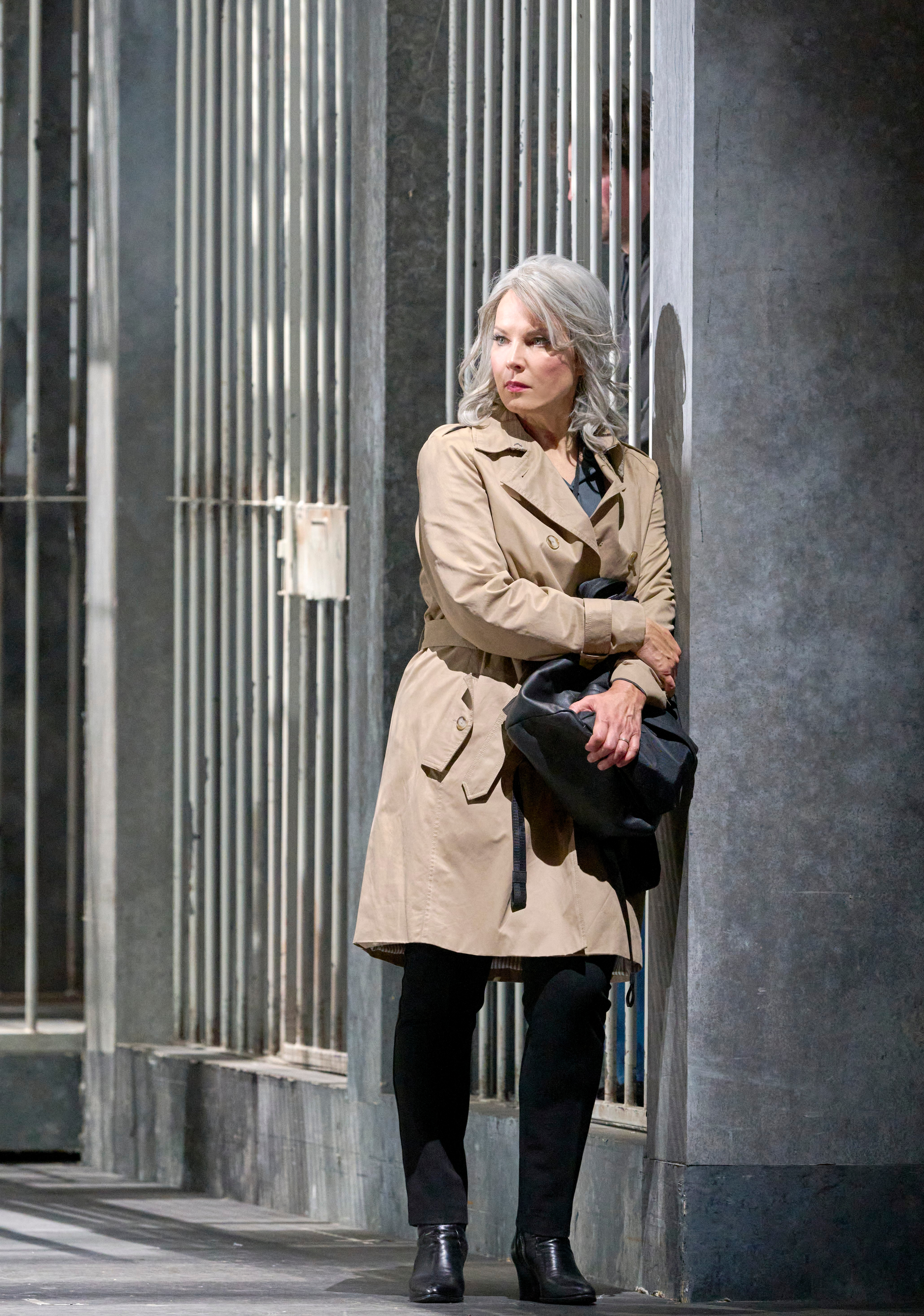 Eine Frau im beigen Trenchcoat steht nachdenklich vor einer Gitterwand und hält eine schwarze Tasche.