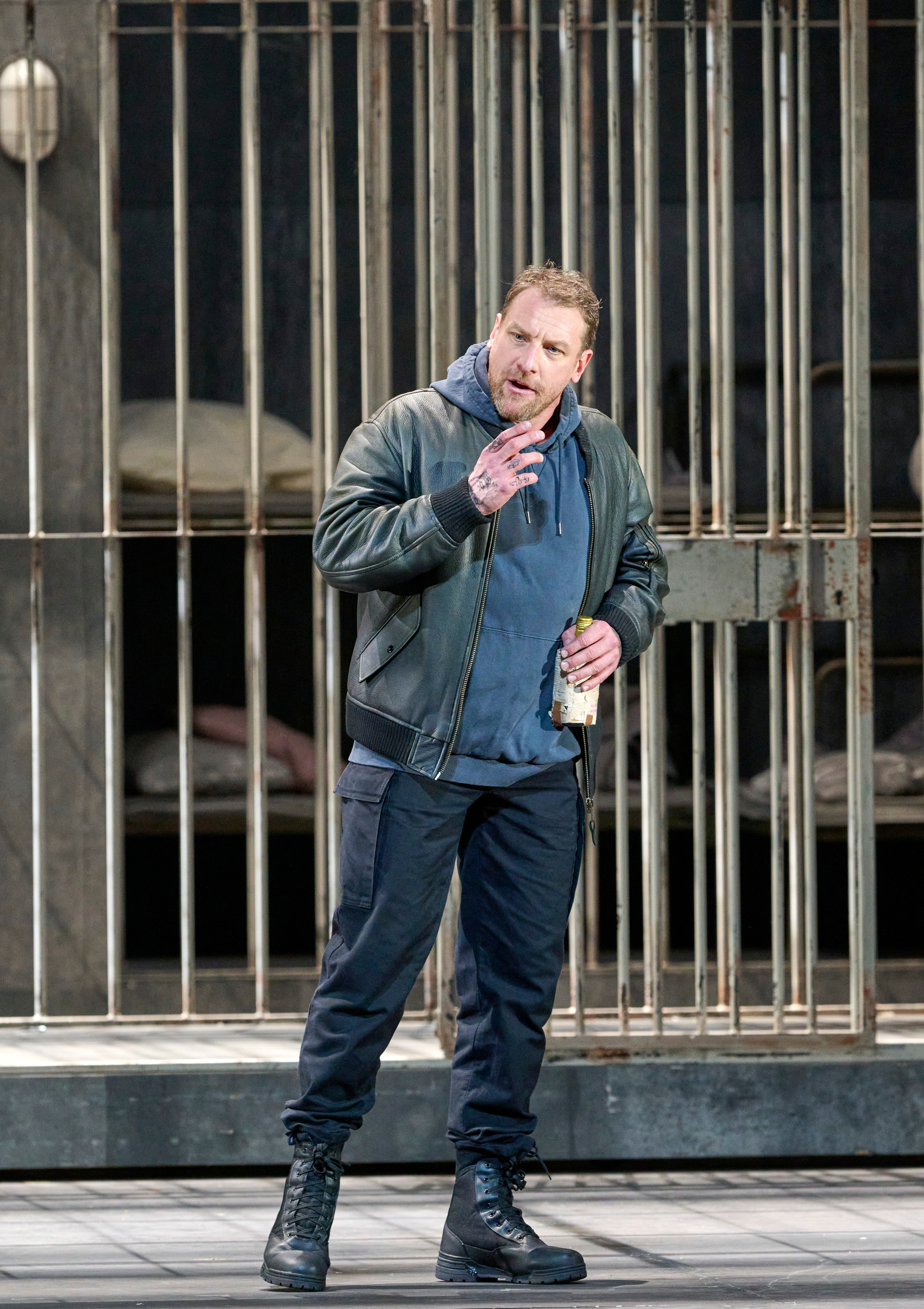 Ein Mann in Freizeitkleidung steht mit einer Bierdose vor einer Gittertür und spricht gestikulierend.