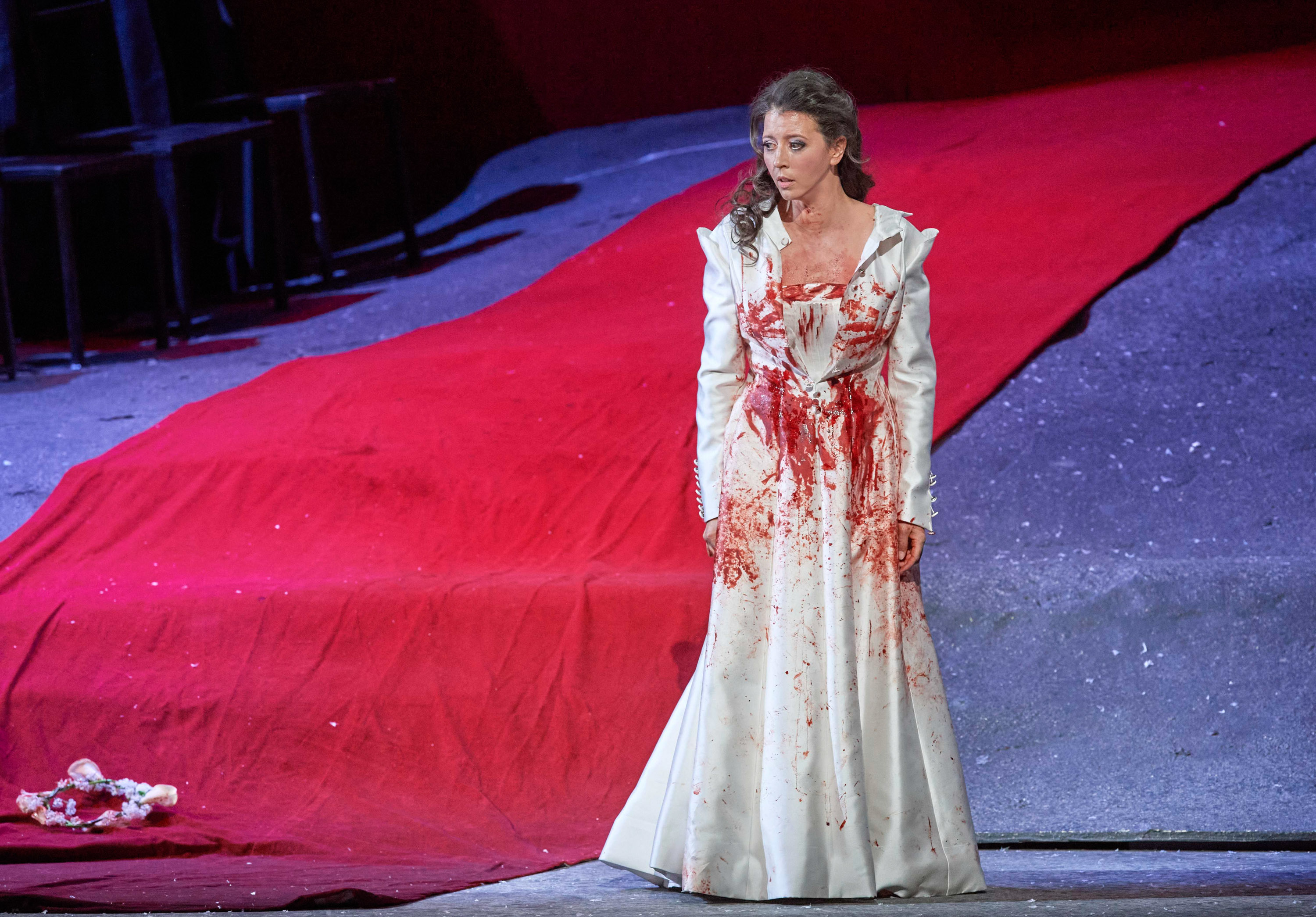 Eine Opernsängerin steht in einem weißen Kleid, das mit roter Farbe oder Kunstblut bedeckt ist, auf einer dramatisch beleuchteten Bühne.