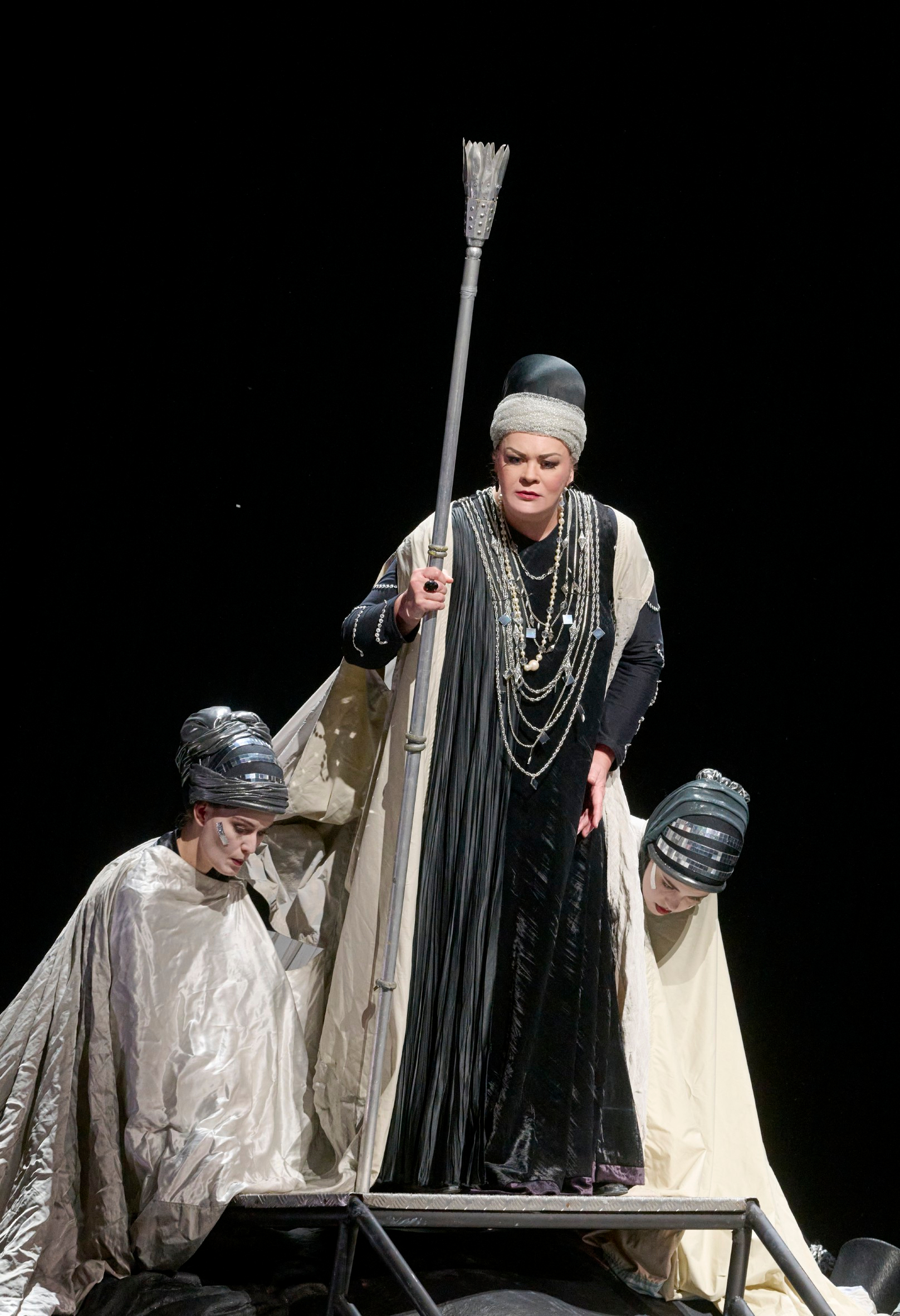 Eine zentrale Figur mit Stab und prunkvollem Kostüm steht auf der Bühne, flankiert von zwei Personen mit silbernen Kopfbedeckungen.