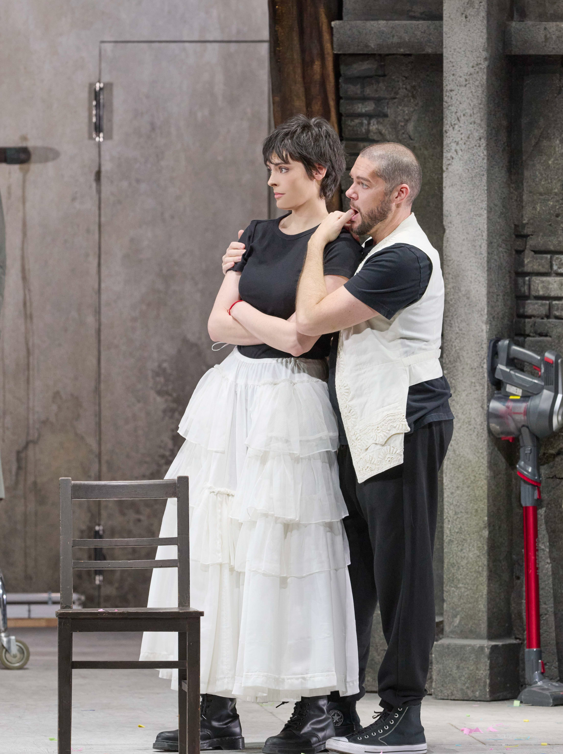 Ein Mann und eine Frau stehen eng umschlungen vor einer grauen Wand; sie trägt ein schwarzes Shirt und einen weißen Stufenrock, er lehnt sich zärtlich an ihre Schulter.