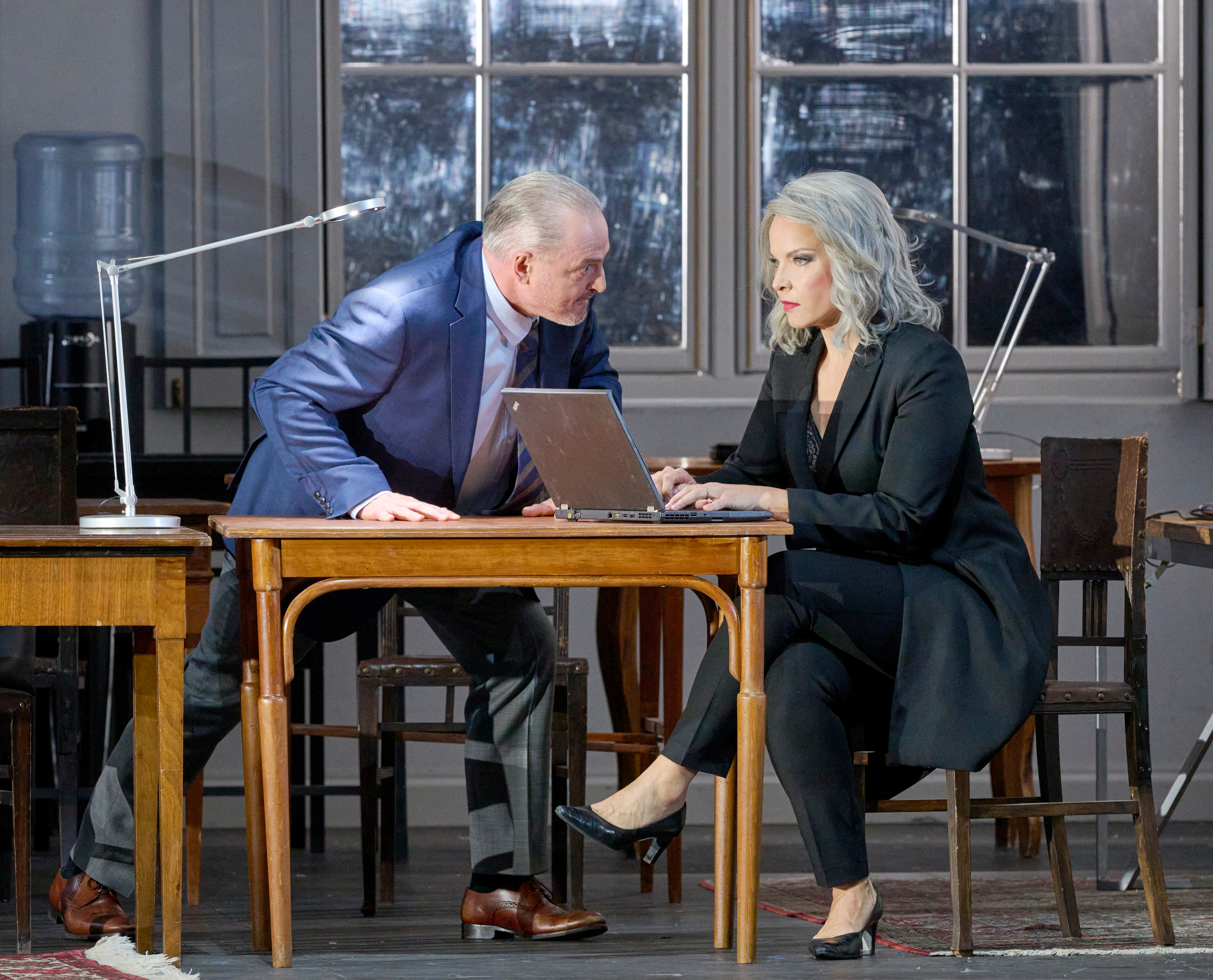 Eine Frau arbeitet an einem Laptop, während ein Mann sich ihr über den Tisch hinweg zuwendet und sie anspricht.