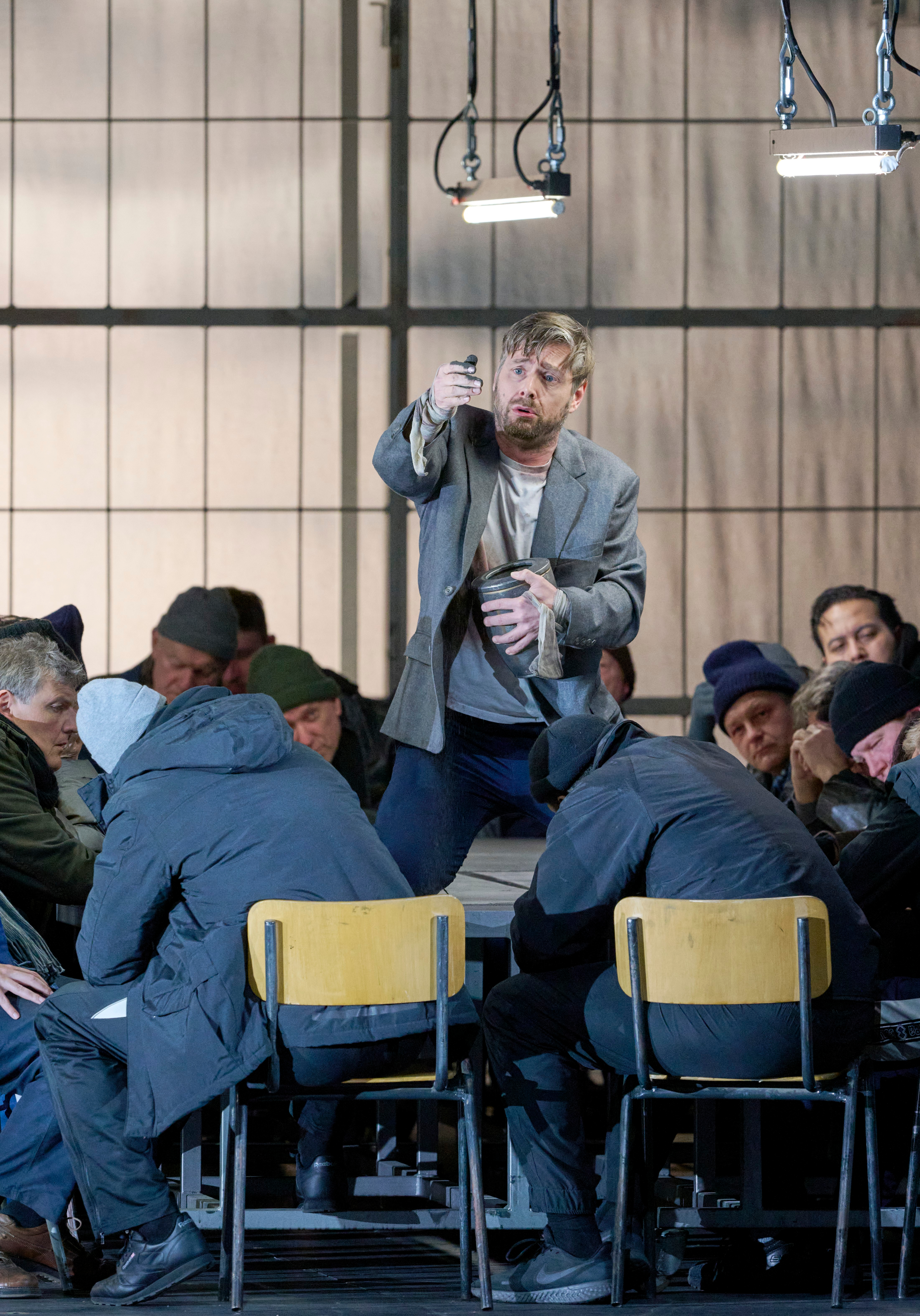 Ein Mann in zerknittertem Anzug steht gestikulierend zwischen mehreren sitzenden Personen in Winterkleidung.