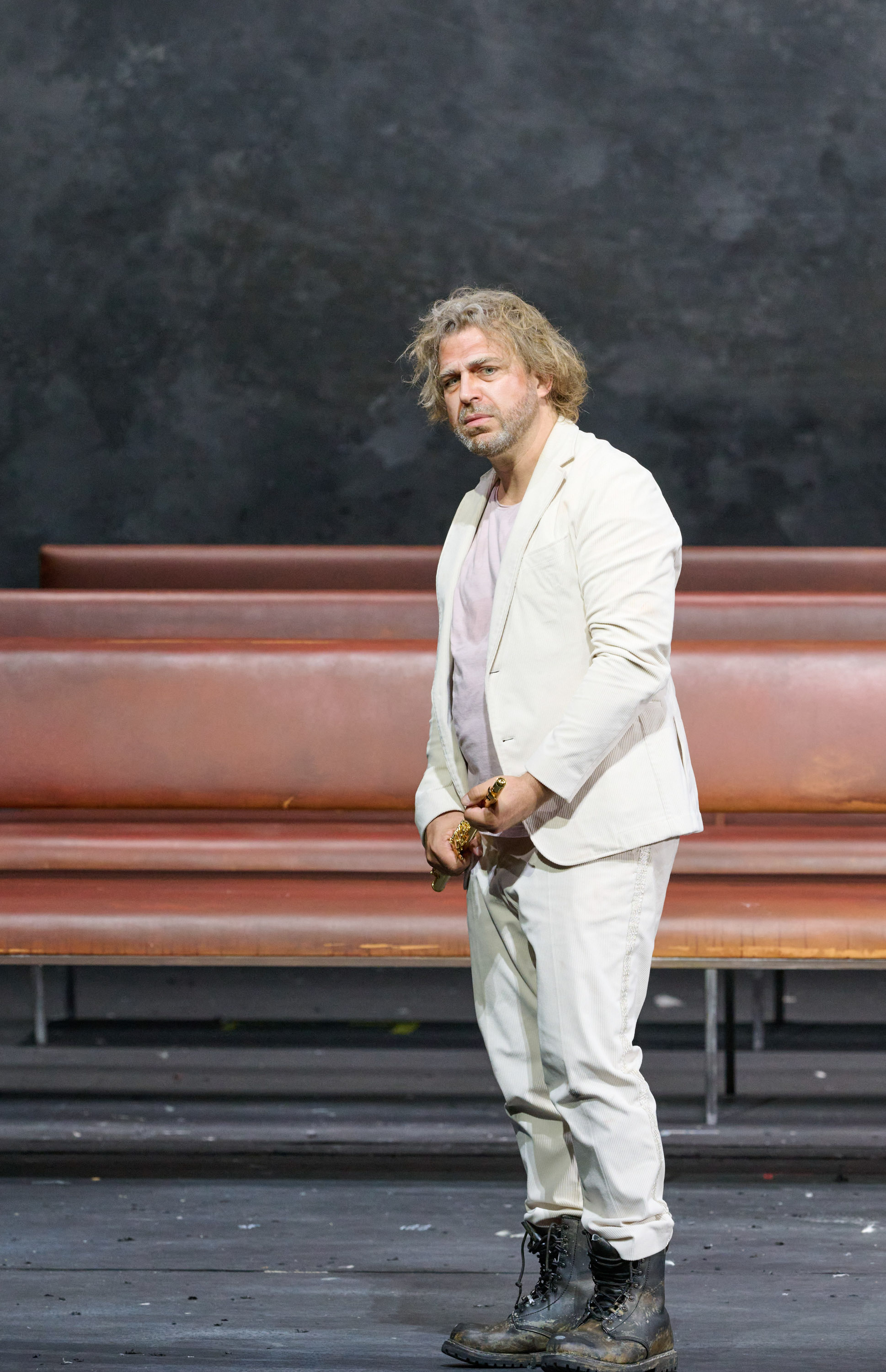 Ein zerzaust aussehender Mann in einem weißen Anzug steht vor einer schwarzen Wand und hält eine goldene Querflöte in der Hand.