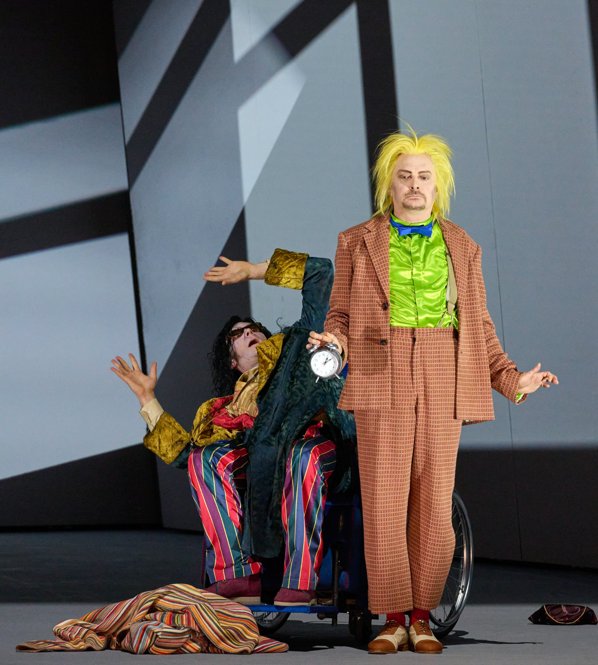 Eine Figur mit gelber Perücke und schrillem Anzug steht vorn, während eine bunt gekleidete Person dramatisch im Rollstuhl im Hintergrund sitzt.