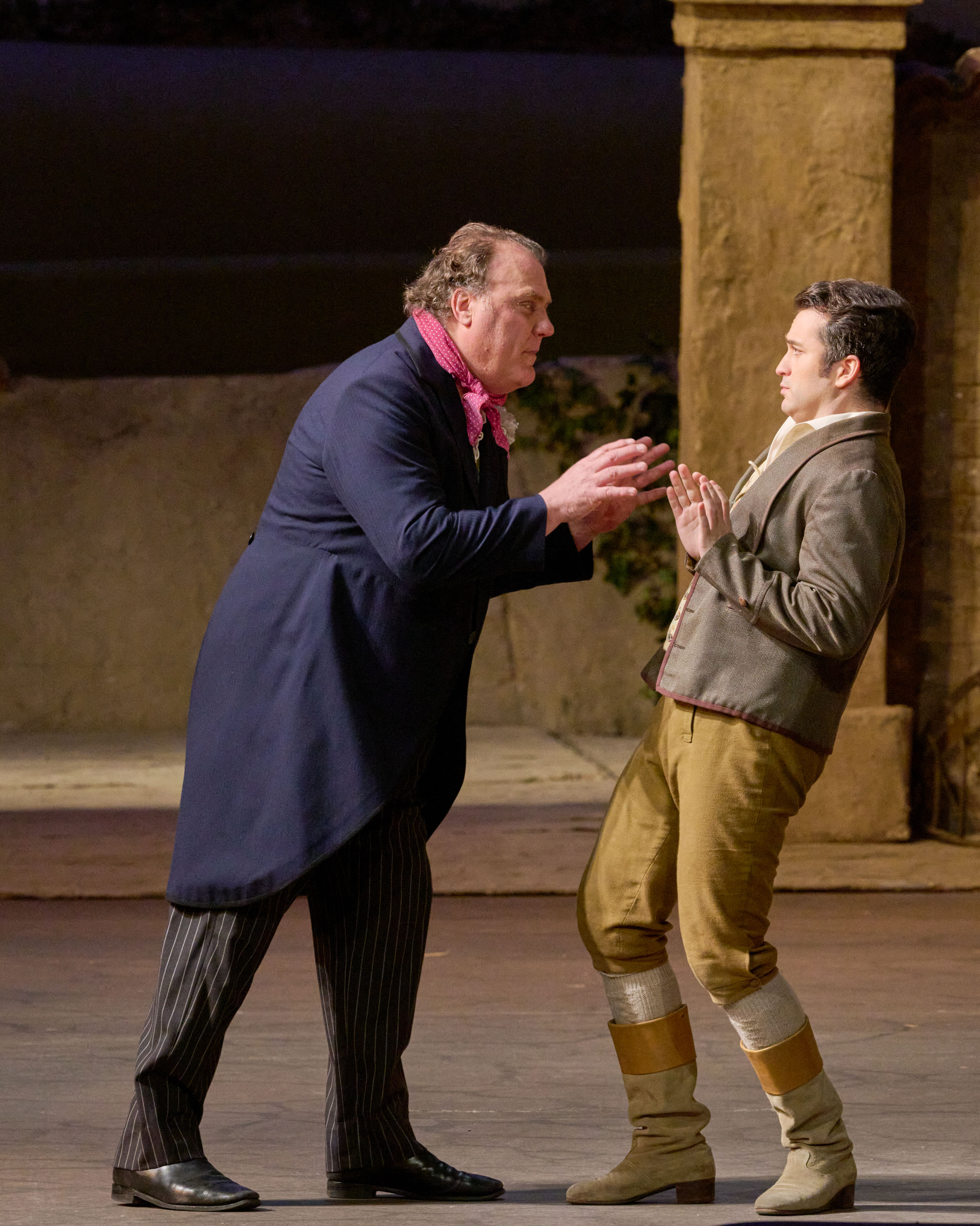 Ein älterer Mann spricht energisch auf einen jüngeren ein, der zurückweicht und überrascht wirkt, beide auf einer Bühnenkulisse.