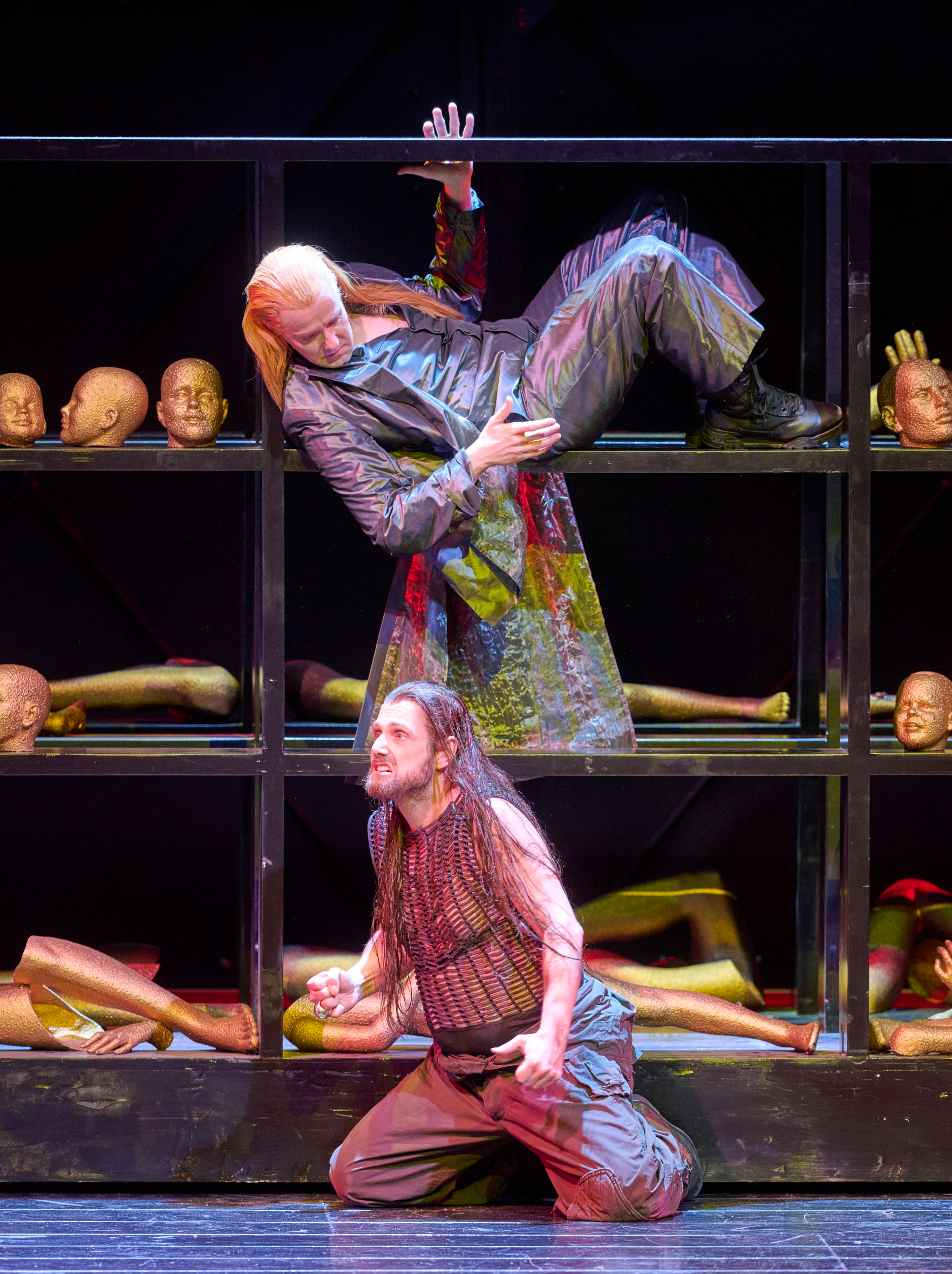 Eine Person in schillerndem Mantel liegt dramatisch in einem Regal voller Köpfe und Gliedmaßen, während eine weitere Figur unten kniet.