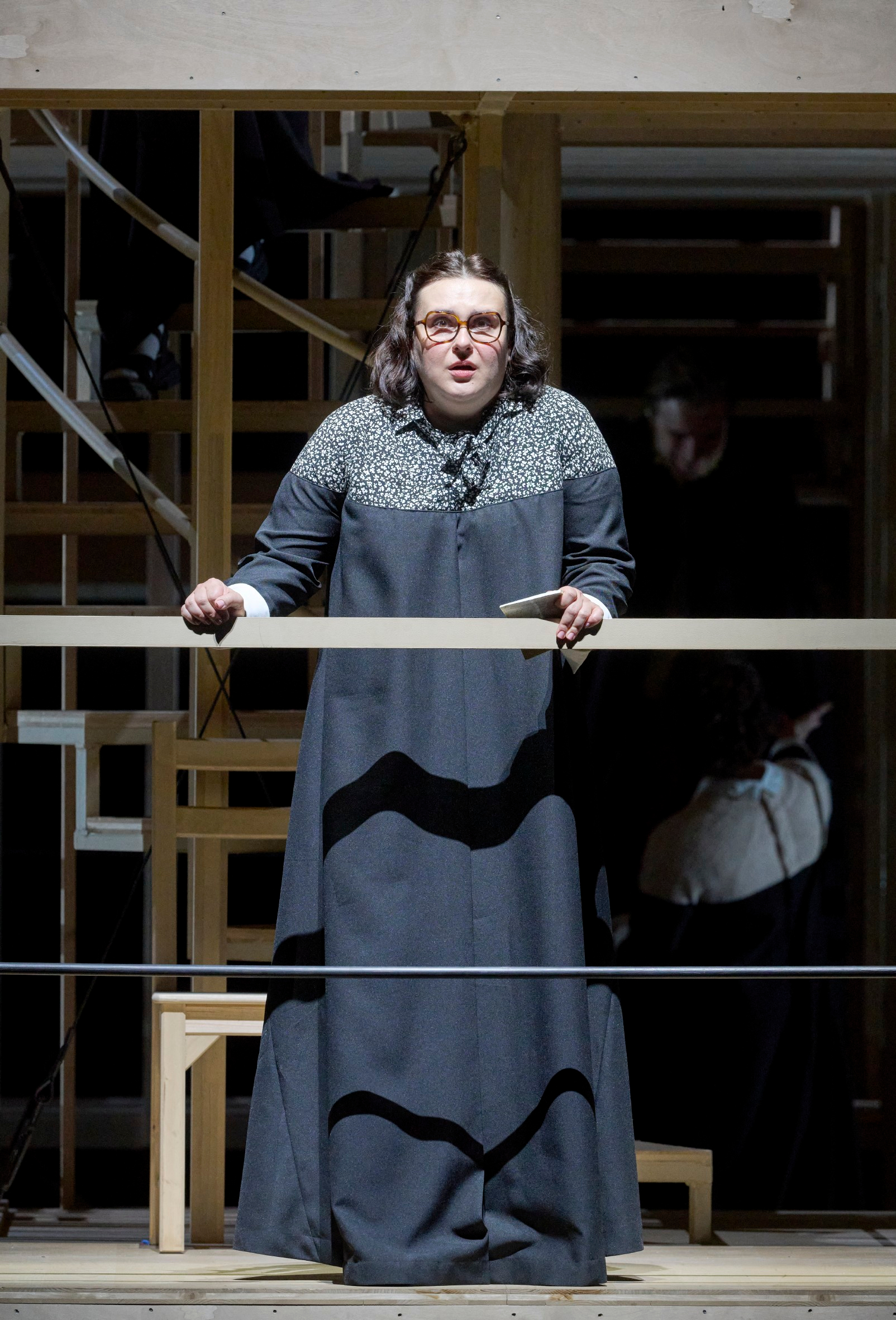 Eine Person mit Brille und mittellangem Haar steht in dunkler Kleidung an einem hölzernen Geländer und blickt direkt nach vorn.