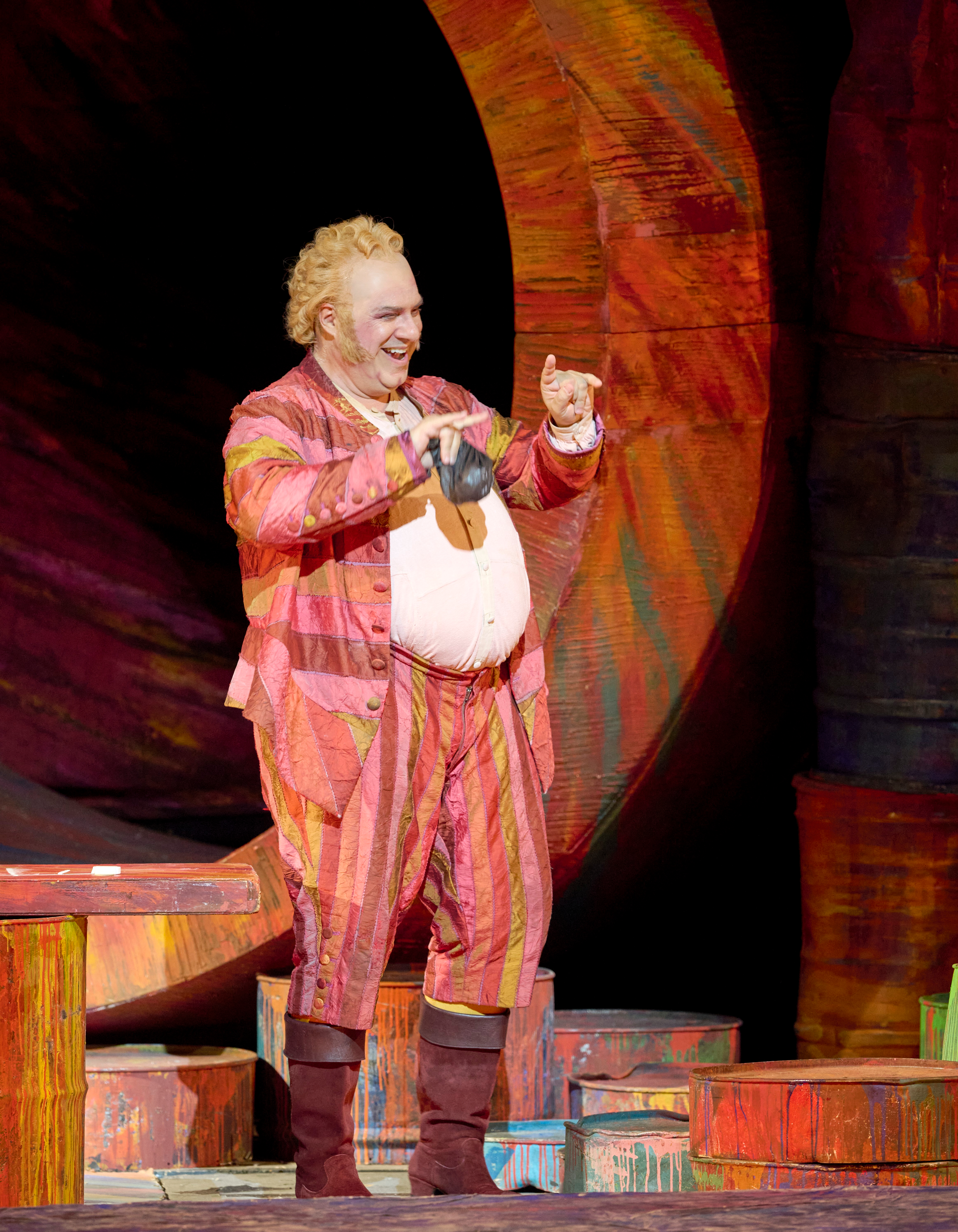 Ein fröhlicher Mann in auffälligem, gestreiftem Kostüm steht lachend auf der Bühne und zeigt mit beiden Händen nach vorn.