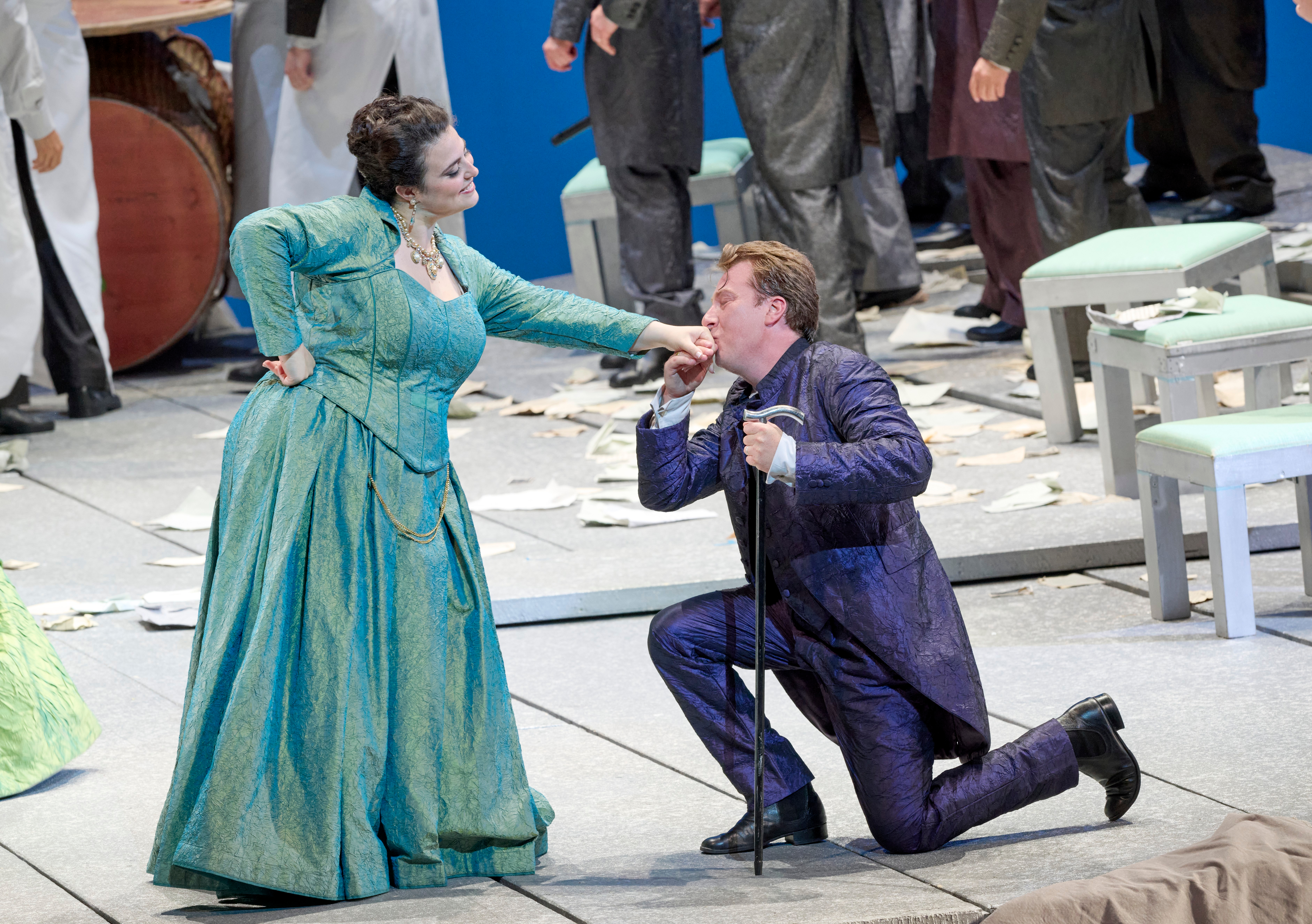 Eine Frau in türkisfarbenem Kleid lässt sich von einem knienden Mann in violettem Anzug galant die Hand küssen.