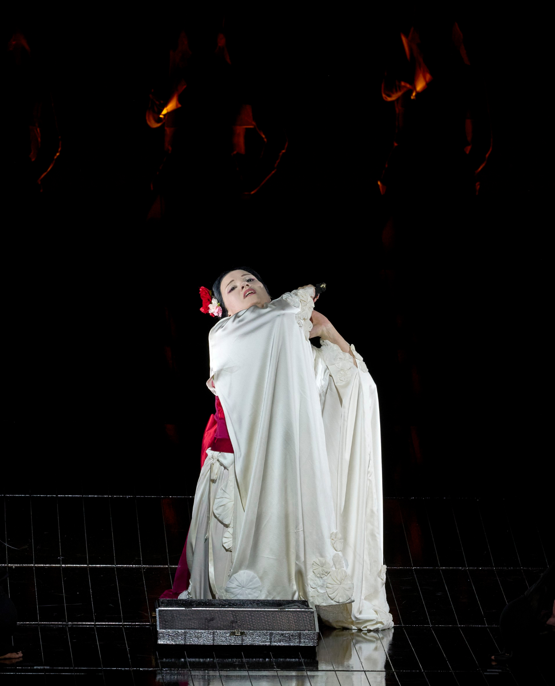 Eine Frau in weißem Gewand mit roter Blume im Haar steht allein im Lichtkegel auf einer dunklen Bühne.