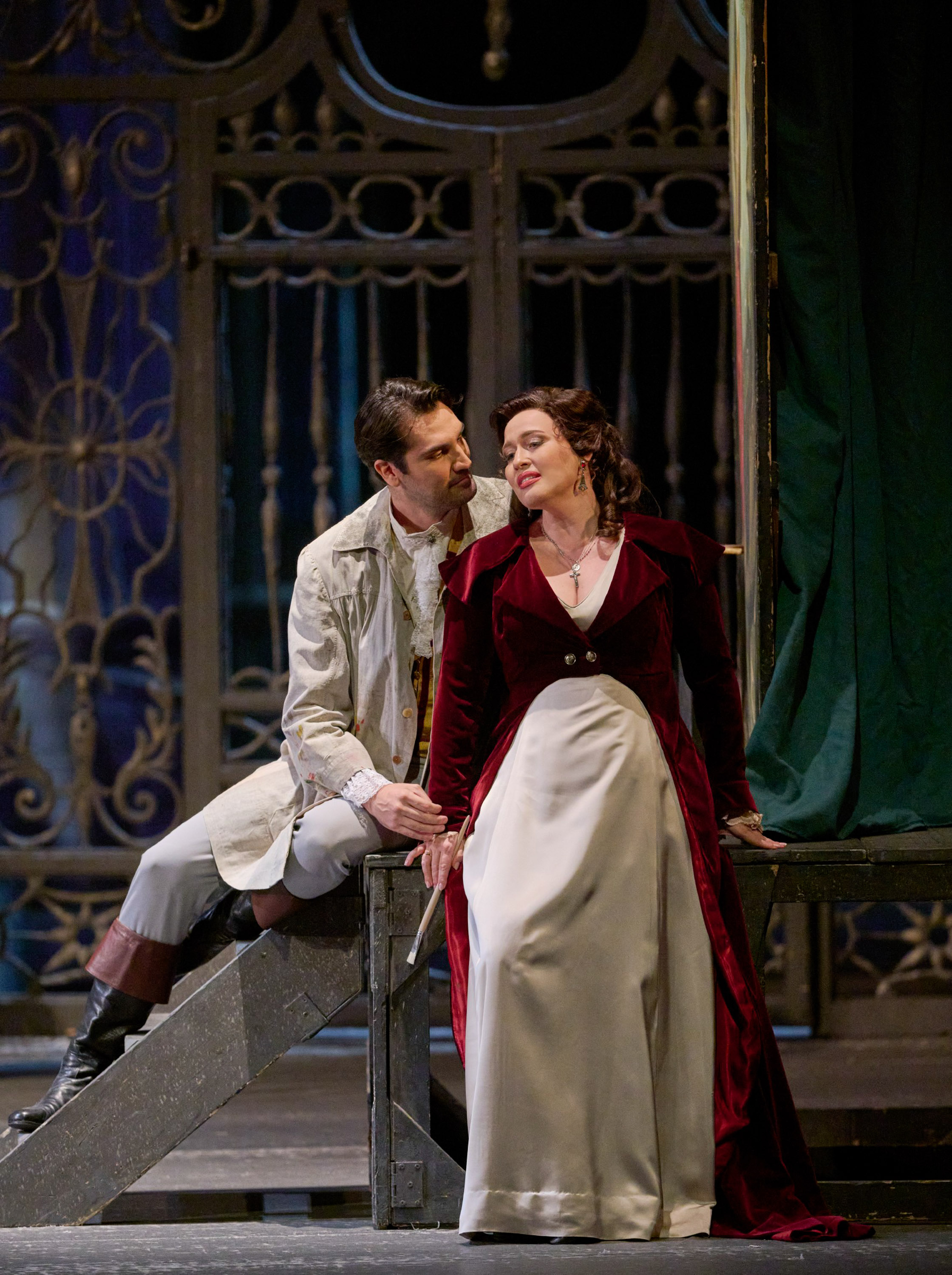 Ein Mann und eine Frau sitzen einander zugewandt auf einer Brücke. Sie trägt einen roten Mantel, er einen weißen.