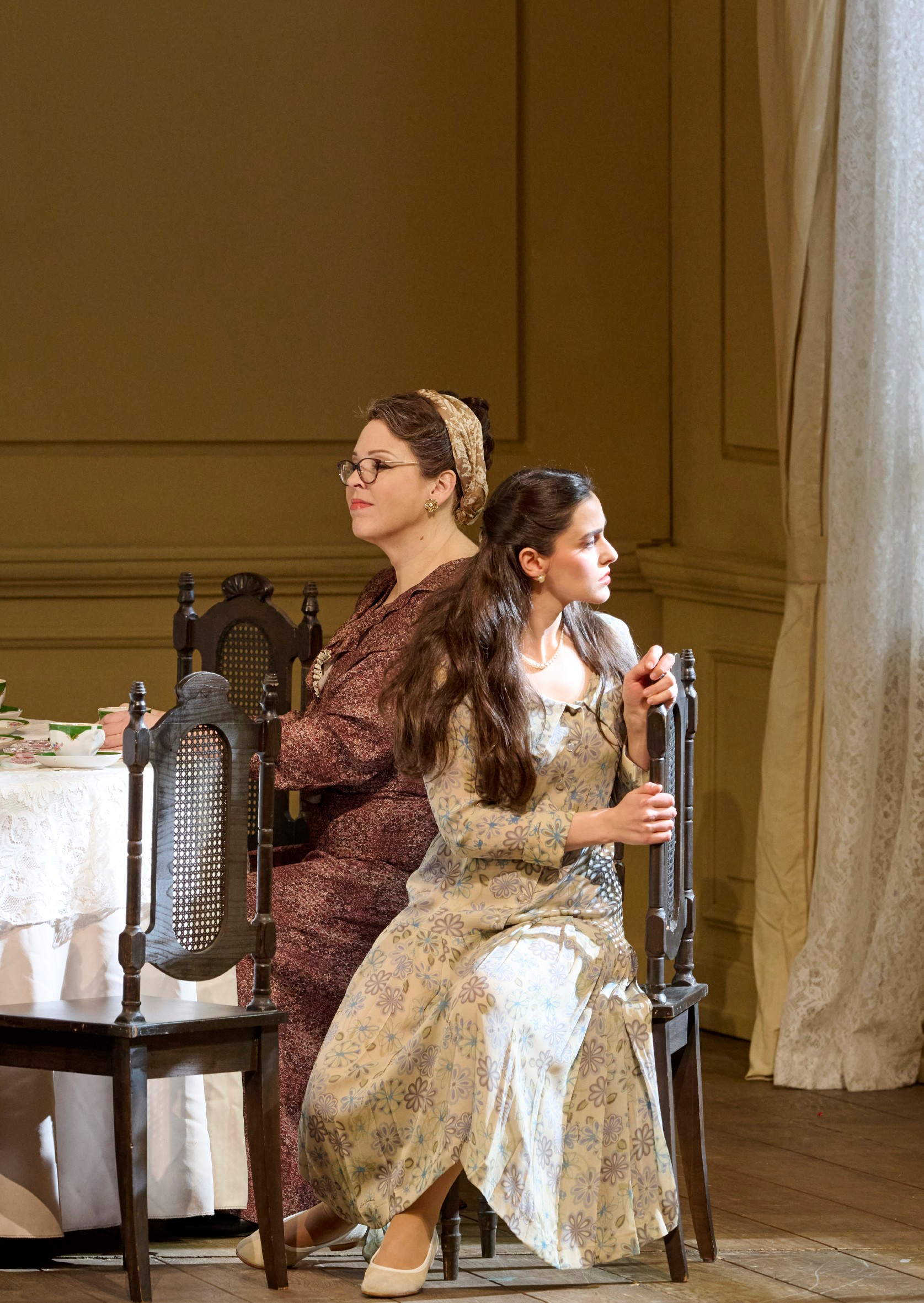 Zwei Frauen sitzen Rücken an Rücken auf Stühlen an einem festlich gedeckten Tisch und blicken in entgegengesetzte Richtungen.