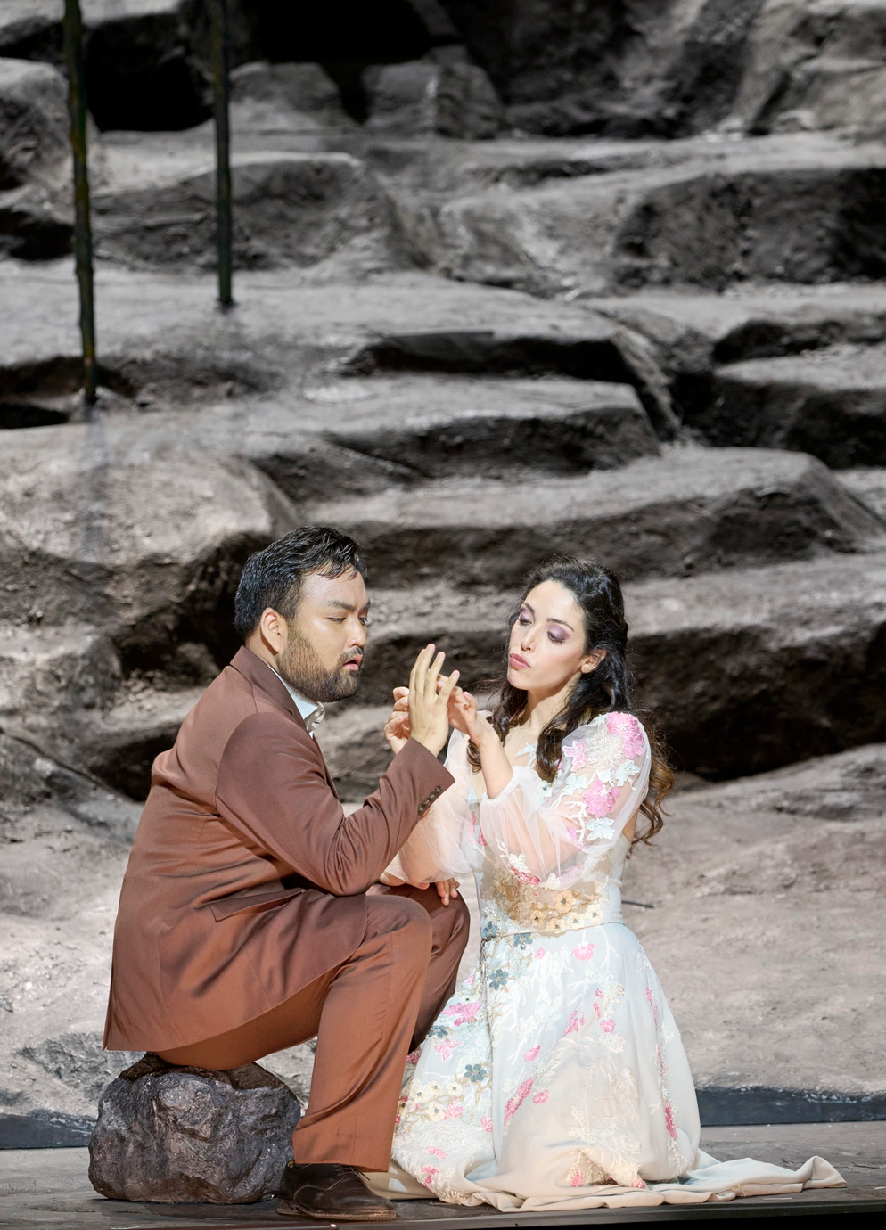 Mann und Frau sitzen auf dem Boden einer Felsenlandschaft; sie halten ihre Hände aneinander.