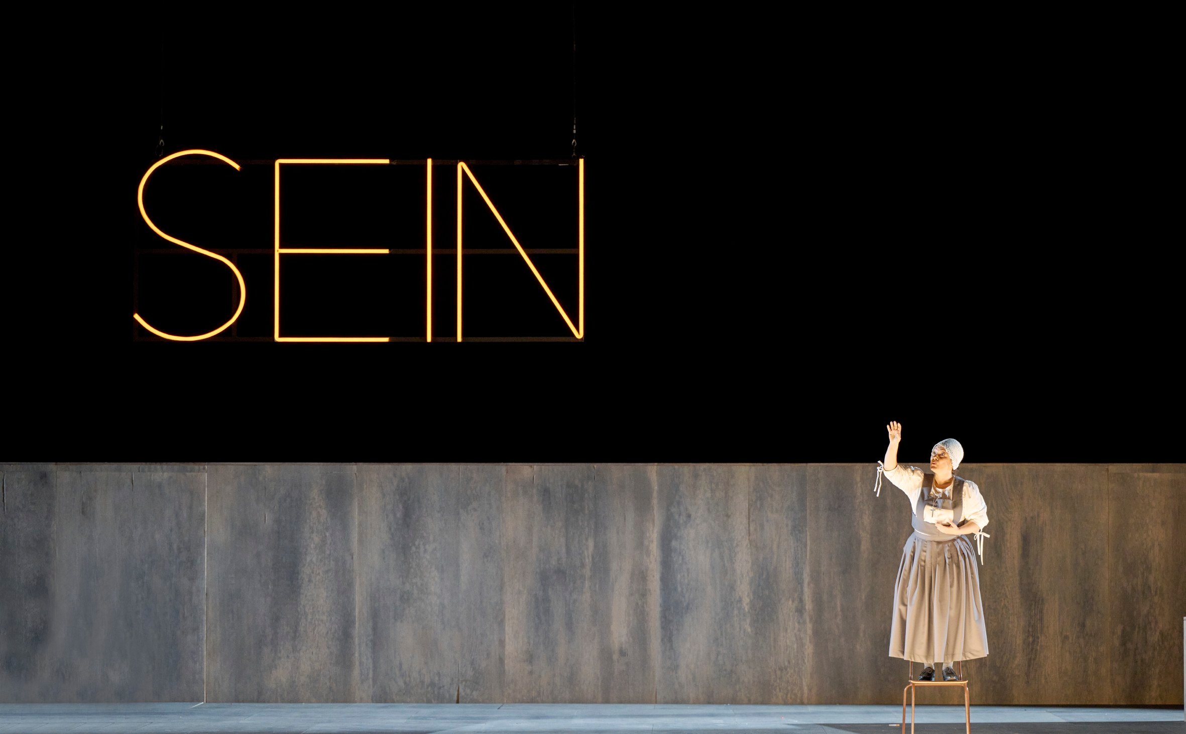 Eine Frau steht vor dunklem Hintergrund; dort ist der Schriftzug "SEIN" zu lesen.