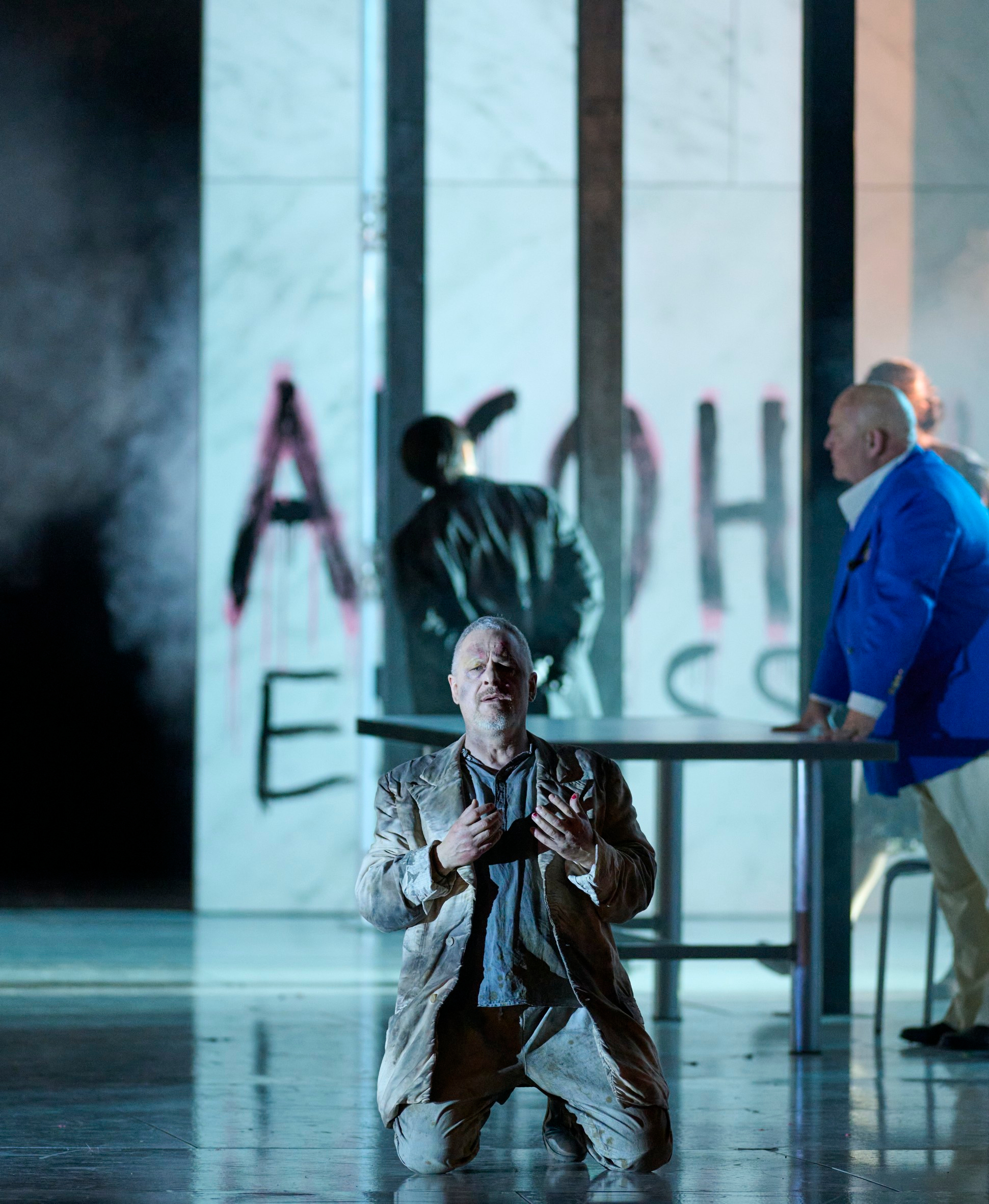 Ein Mann kniet verzweifelt auf der Bühne, während zwei weitere Figuren in modernem Outfit an einem Tisch stehen; im Hintergrund ist „ALL ANIMALS ARE EQUAL“ gesprüht.